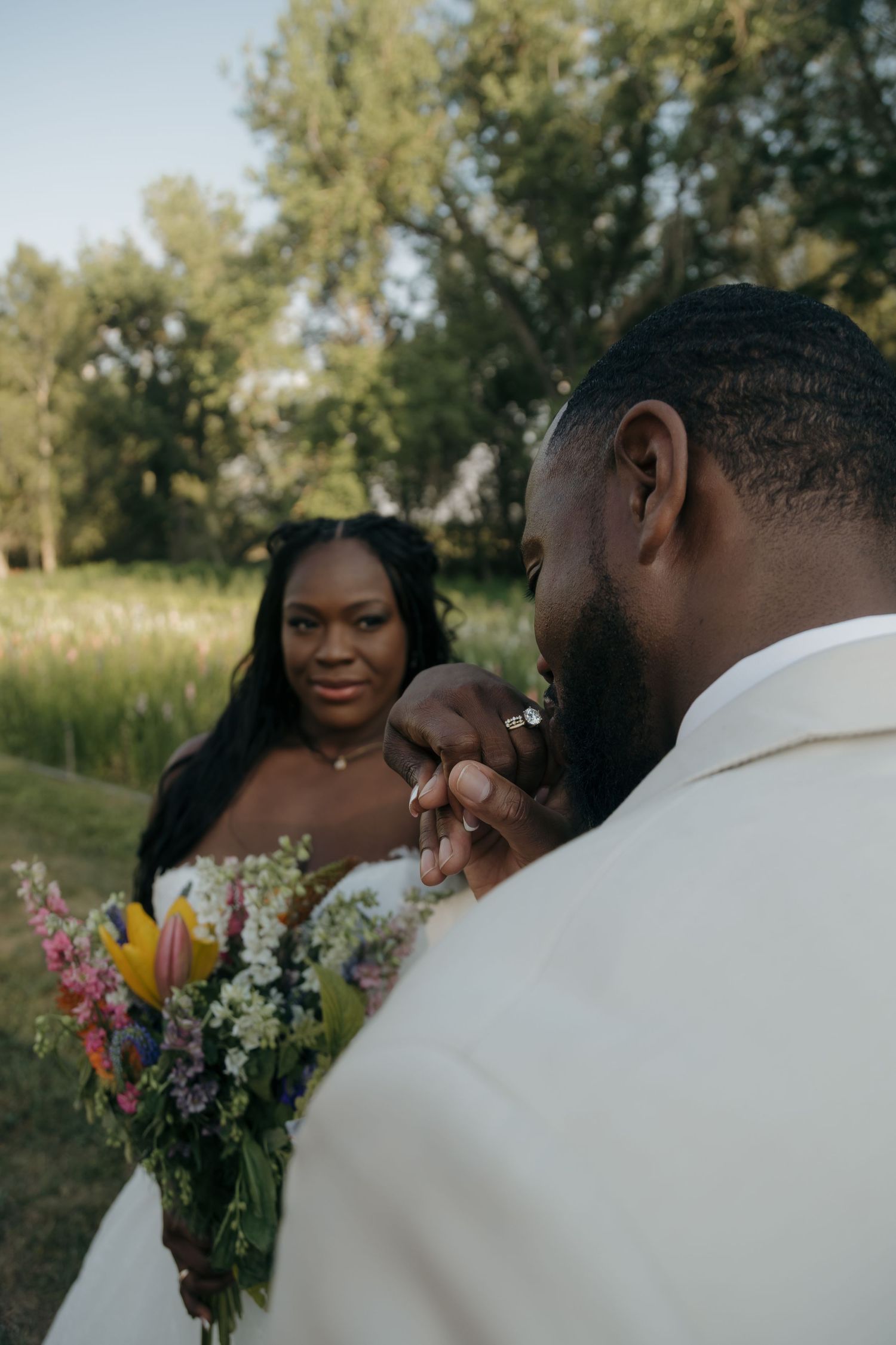 Groom holds bride's hand with engagement ring visible during outdoor wedding ceremony with colorful bouquet.