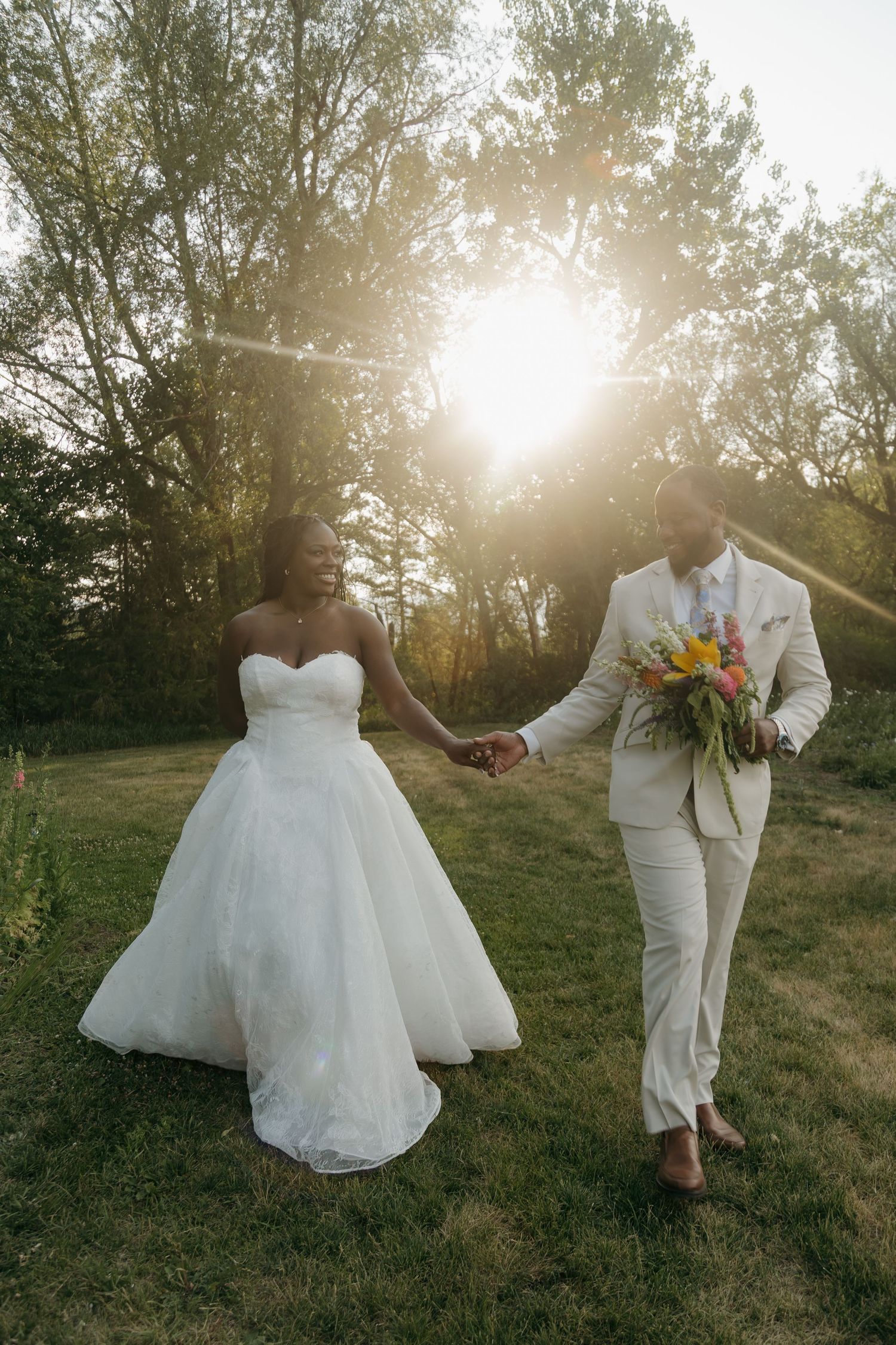 A bride and groom walk hand-in-hand outdoors at golden hour, she in a white gown, he in a cream suit holding a bouquet.