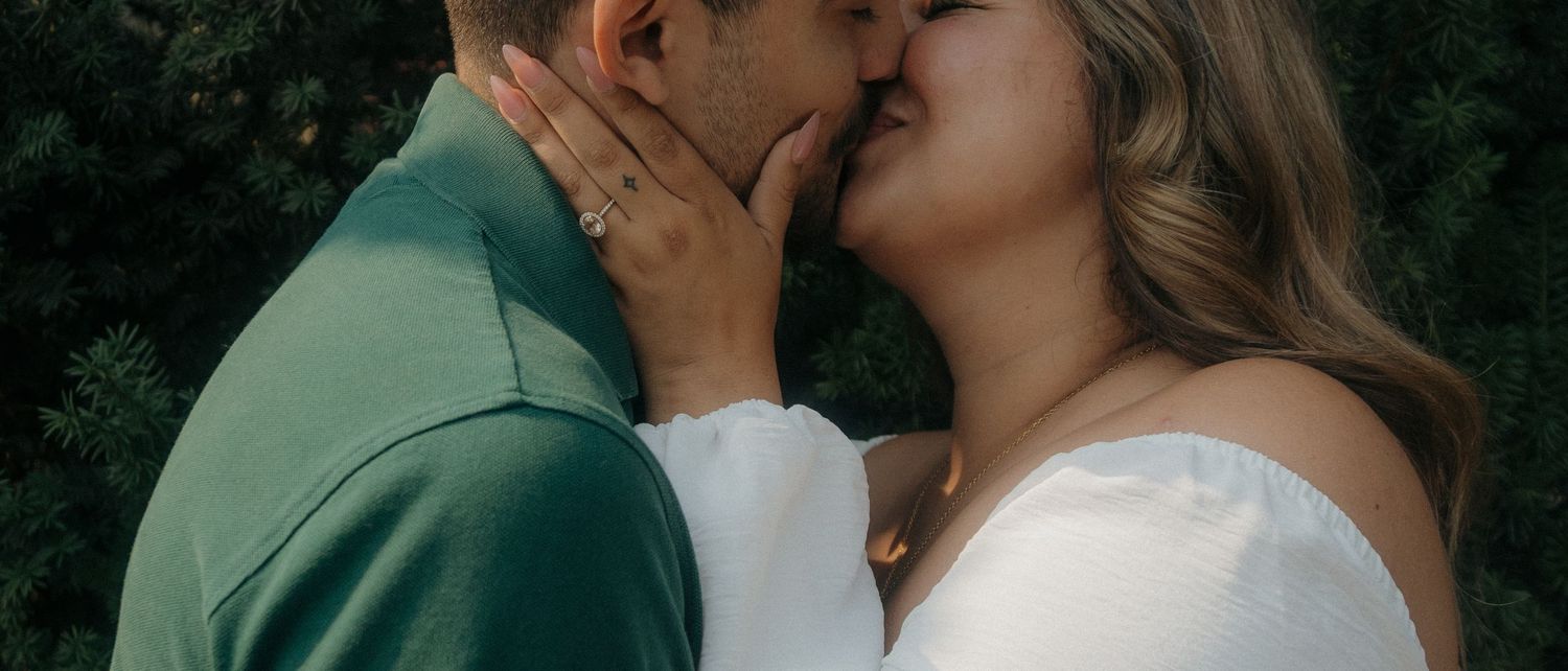 A couple shares a romantic kiss outdoors, with the woman in a white dress and man in a green shirt surrounded by lush greenery.