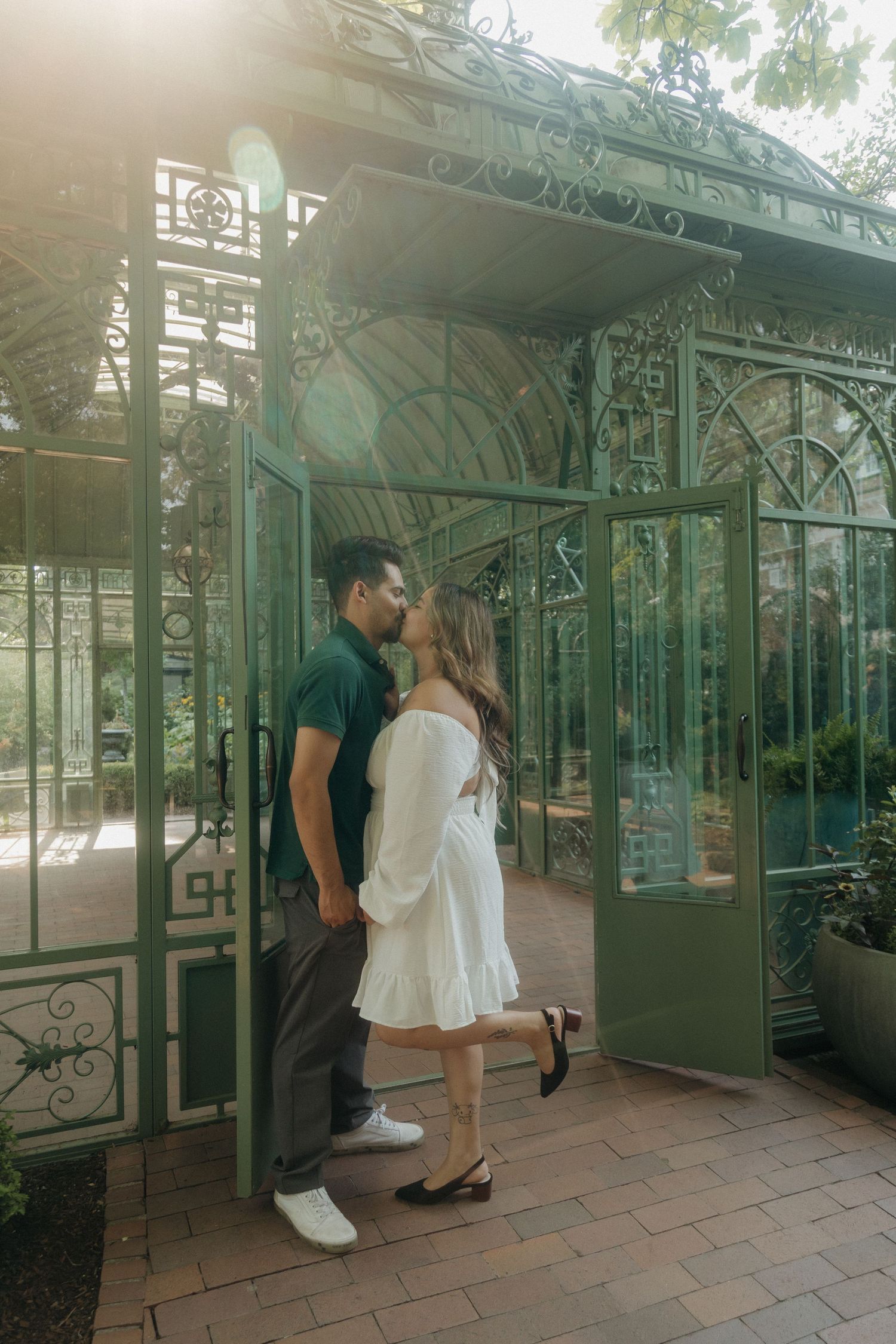 Couple kissing at entrance of ornate green iron gazebo in sunlit botanical garden setting.