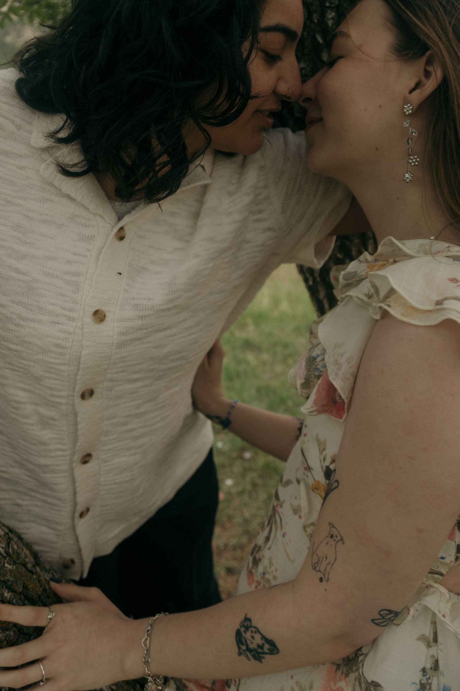 Two women share a romantic almost-kiss outdoors, one in a white button-up and one in a floral dress with tattoos.