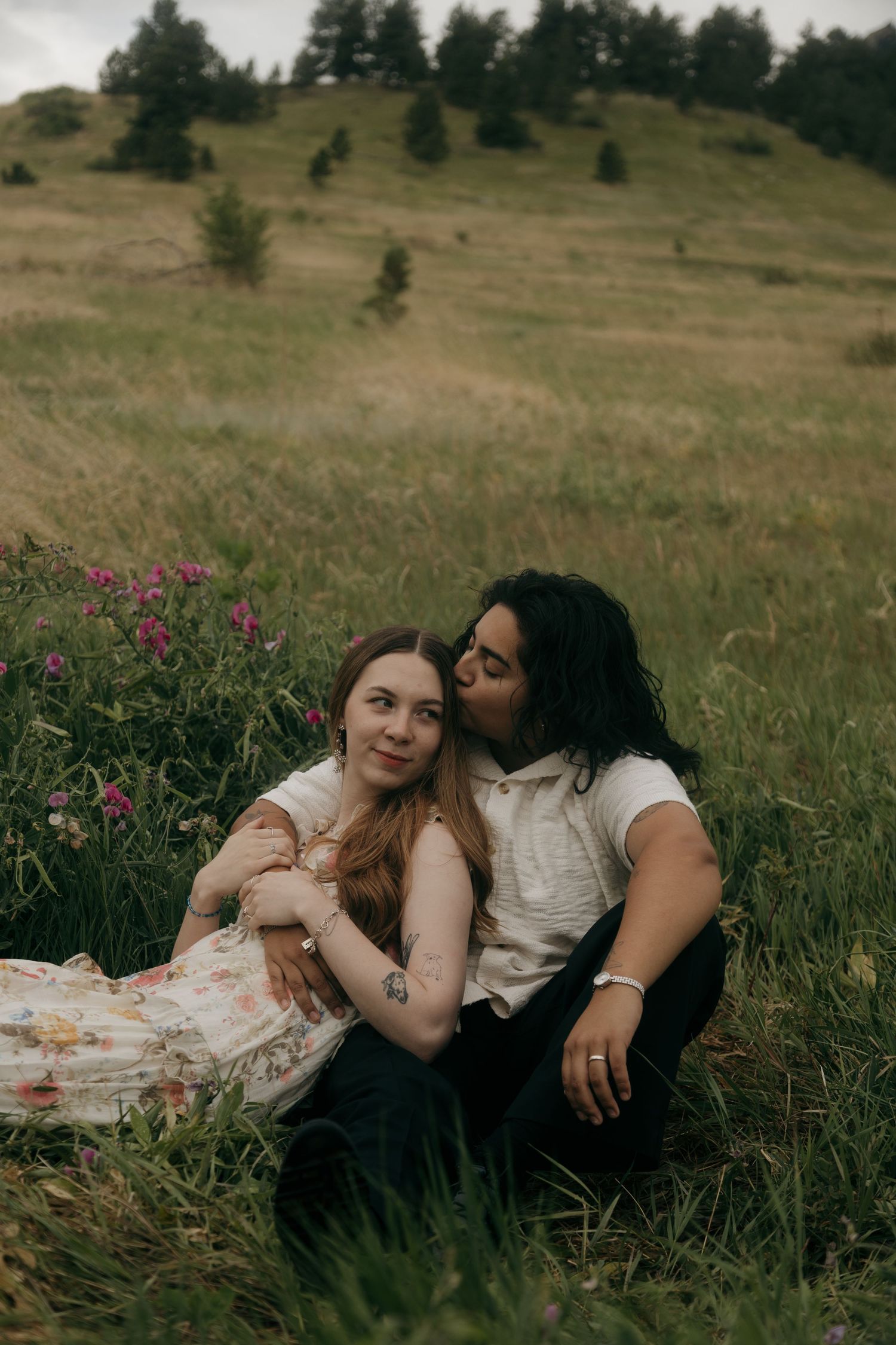 Couple sitting in green meadow with wildflowers, woman in floral dress leaning back as partner kisses her cheek.