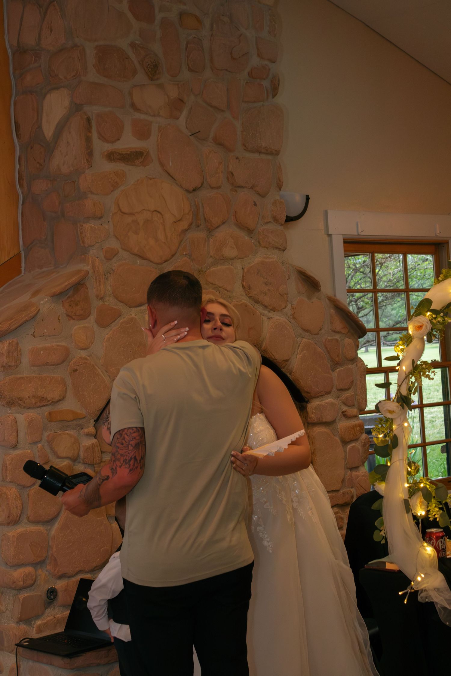 Intimate wedding moment captured in front of rustic stone fireplace with decorative lights.