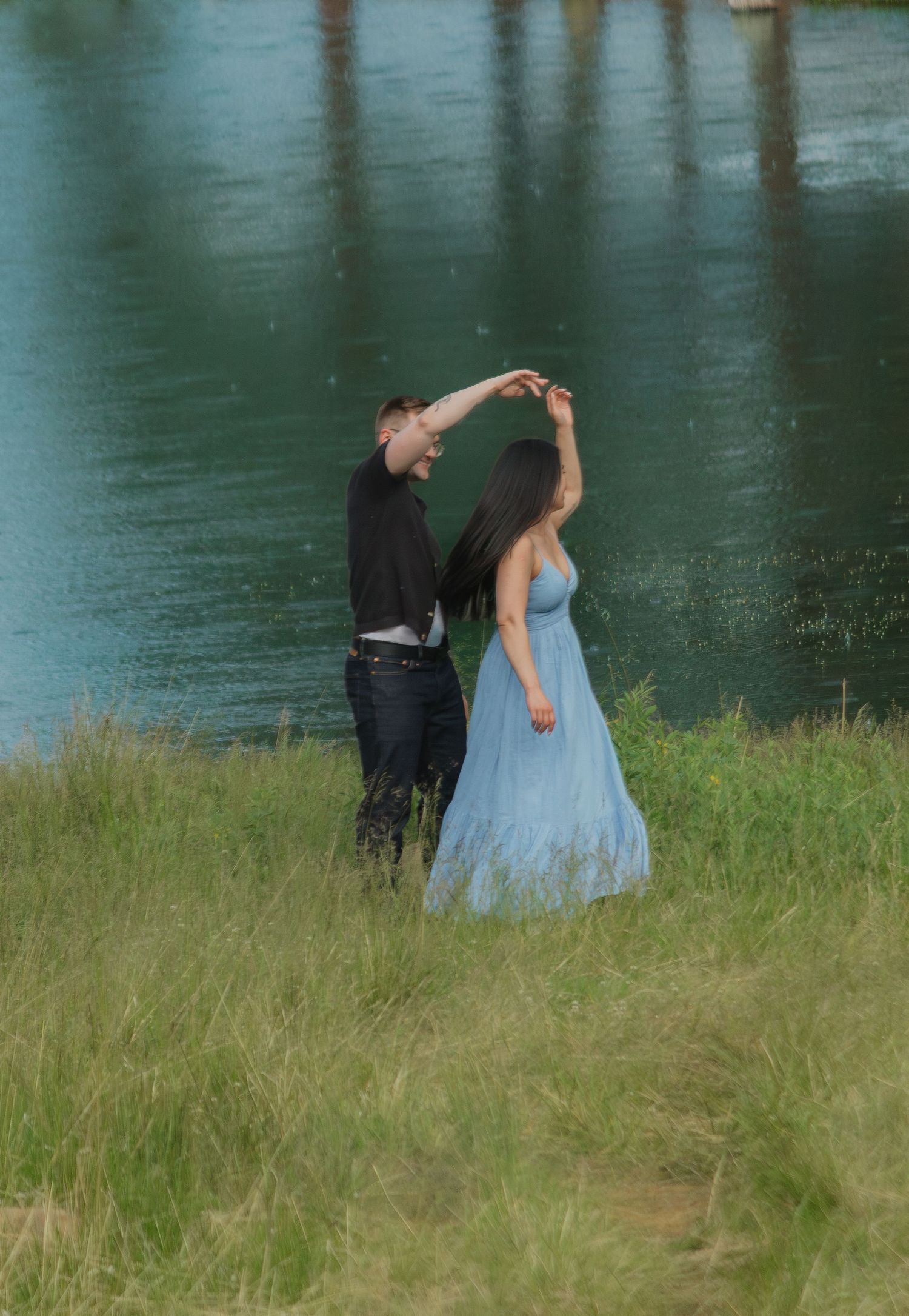 Couple in formal wear sharing a romantic moment by a serene lake with green grass in the foreground.