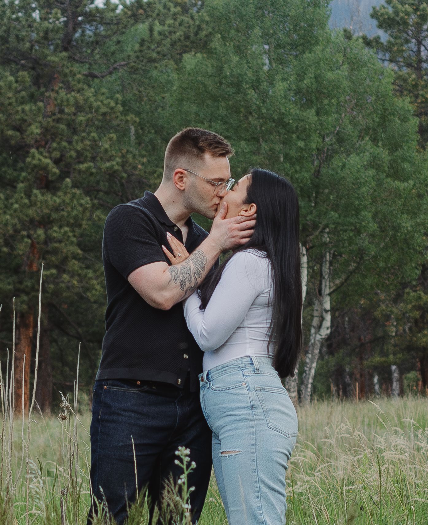 A romantic couple shares a kiss in a mountain field with pine trees and mountains behind them.