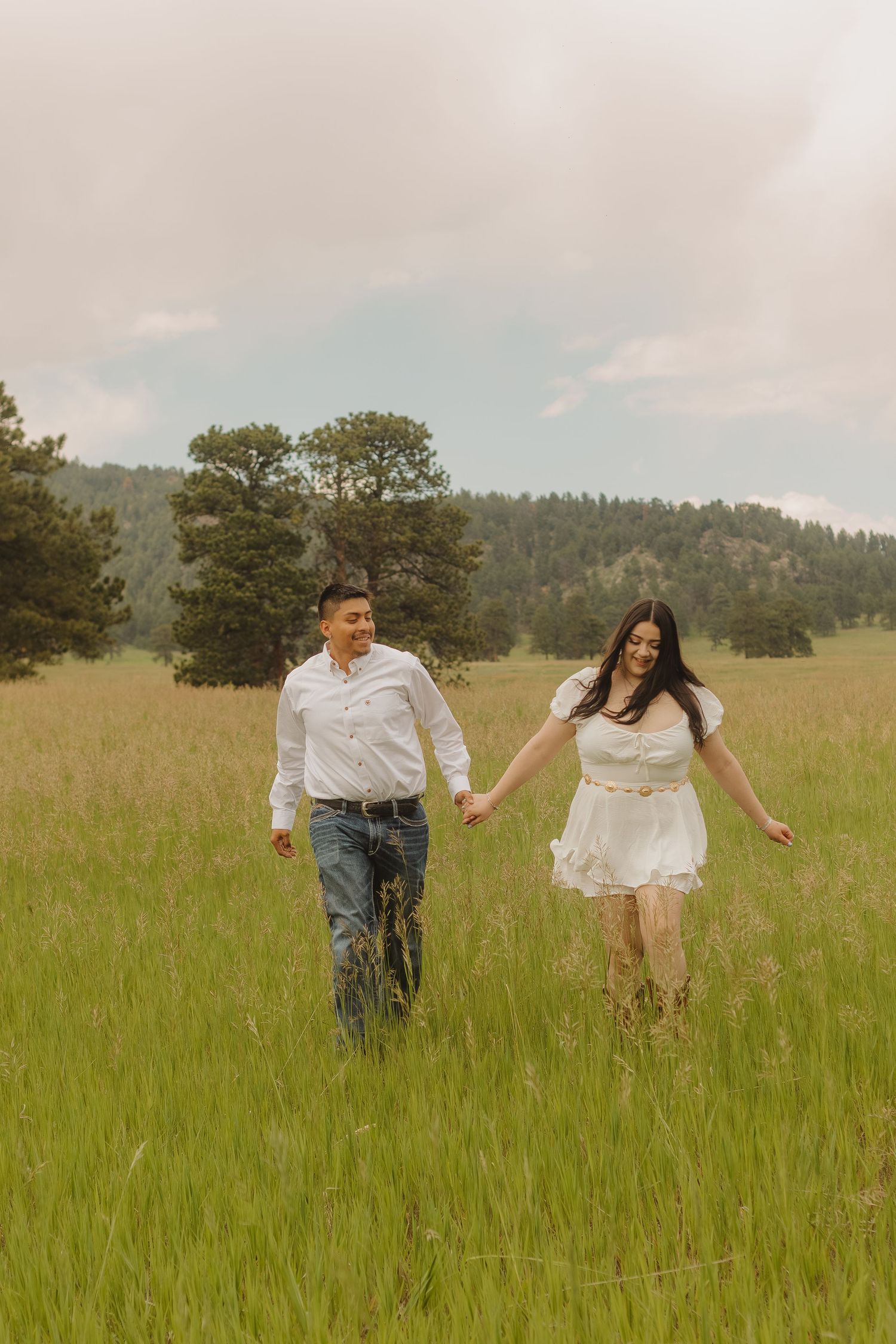 Couple running hand in hand through a sunny meadow with mountains in background.