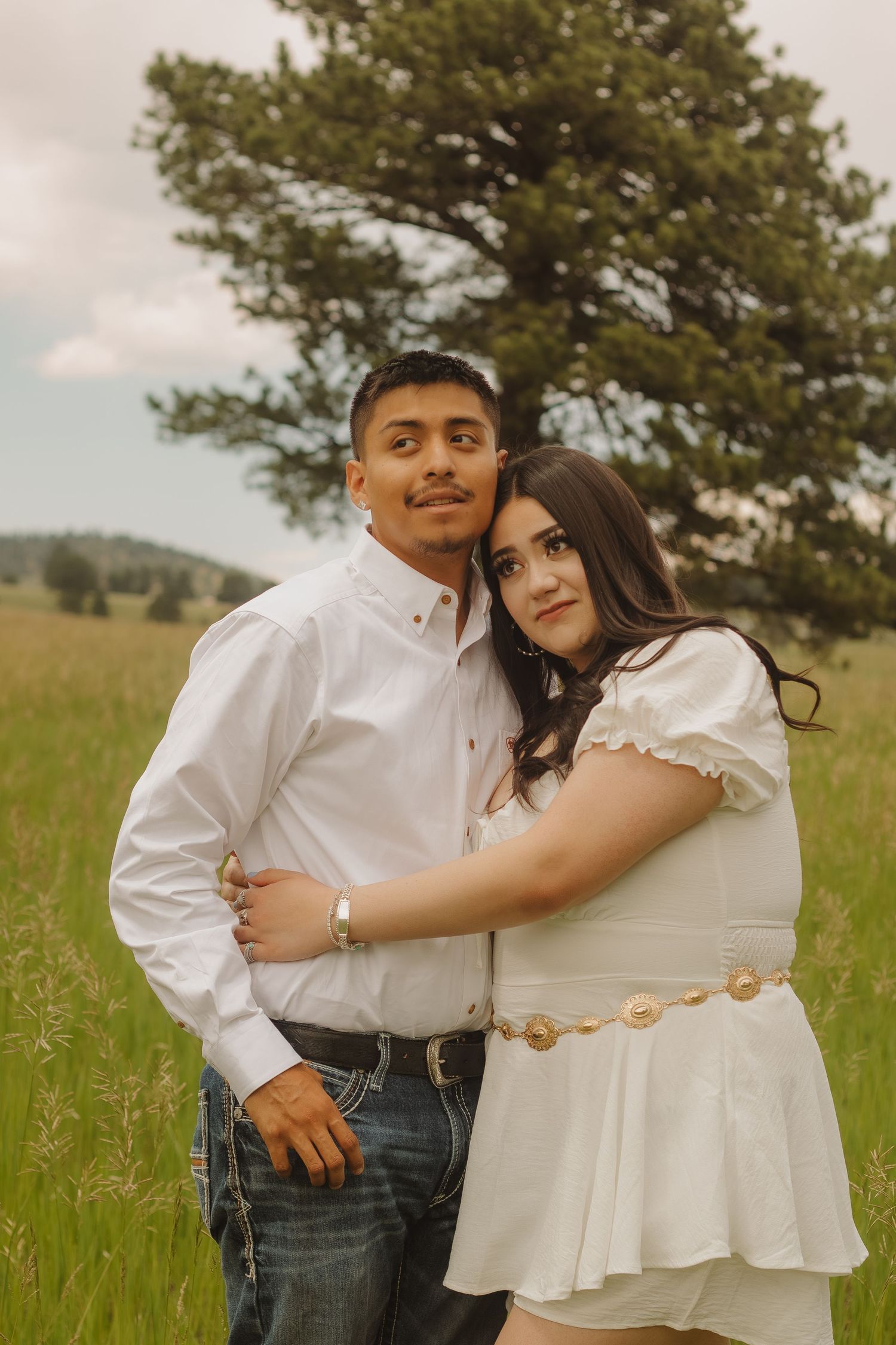 Couple in coordinating white outfits embrace in front of a pine tree in a mountain meadow.
