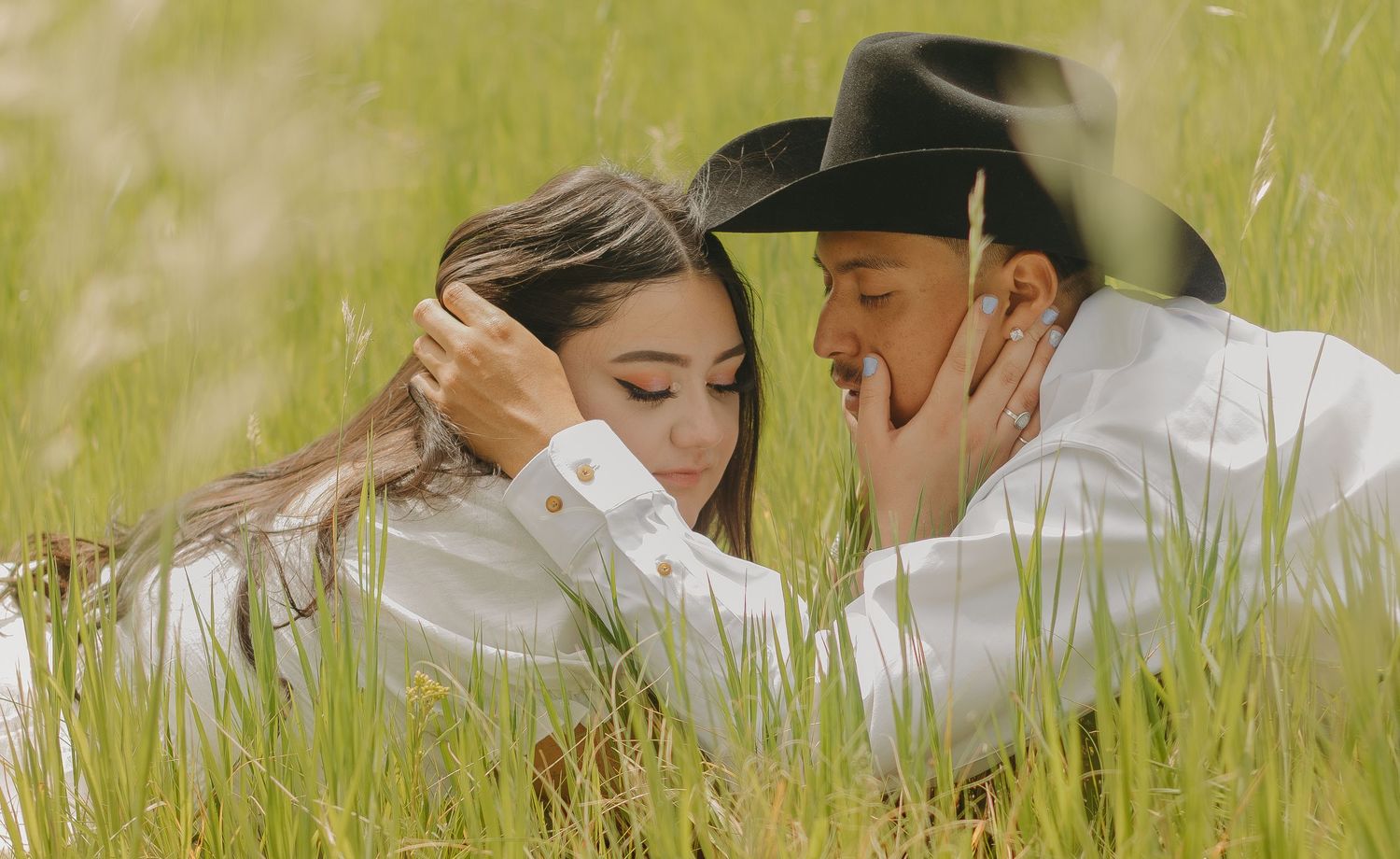 A romantic couple lying in tall grass with one wearing a cowboy hat shares an intimate moment in a field.