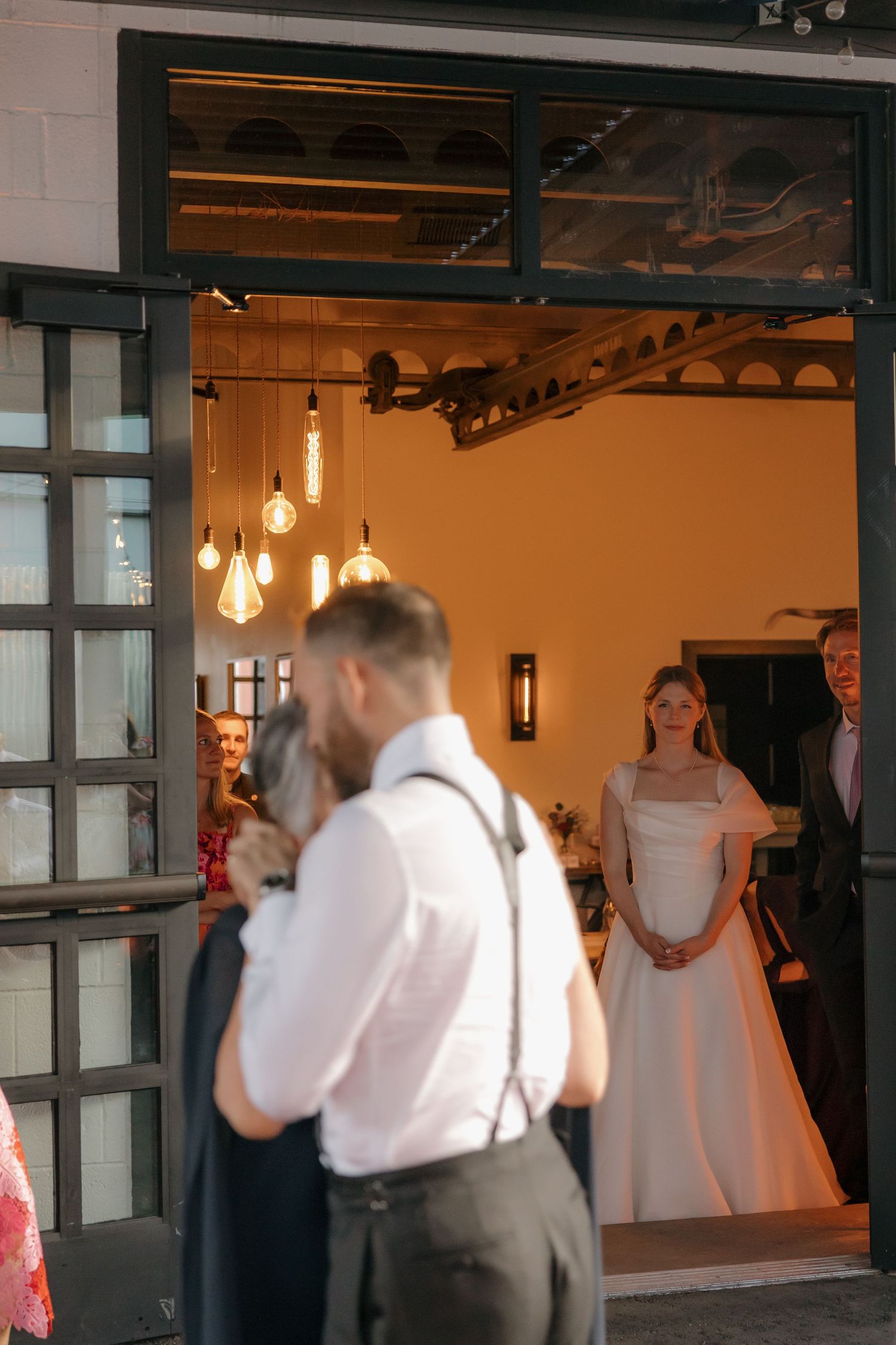 Bride in white gown standing in warmly lit venue doorway as wedding guests gather outside.