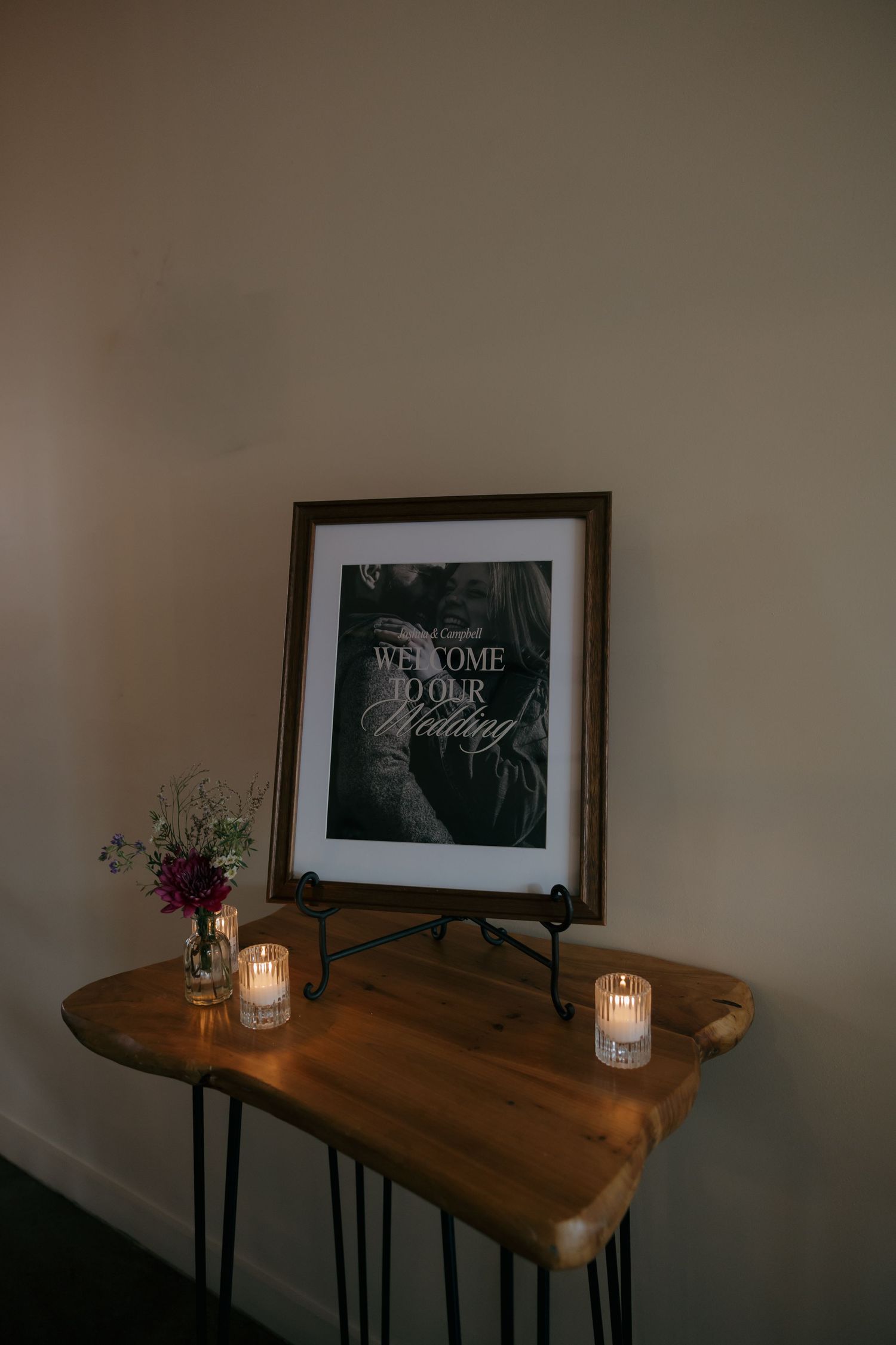 Rustic wooden table with welcome wedding sign in dark frame, candles, and small floral arrangement.
