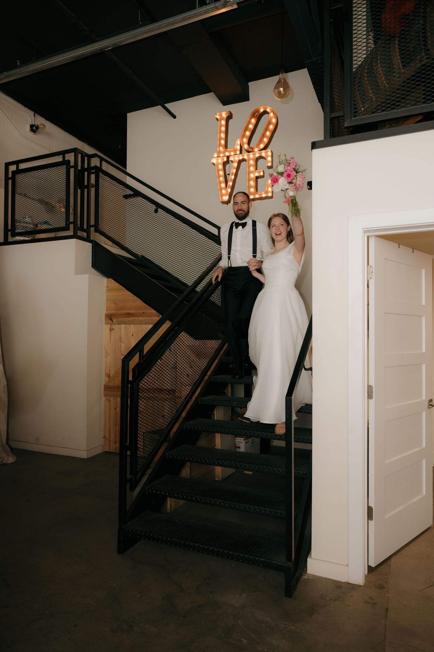 Bride and groom descend industrial staircase beneath glowing marquee LOVE sign at wedding reception venue.
