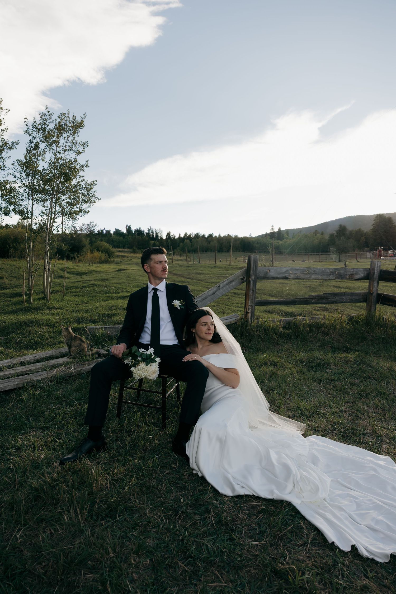 Bride and groom sit on chairs in a green meadow with mountain views, wearing wedding attire on their special day.