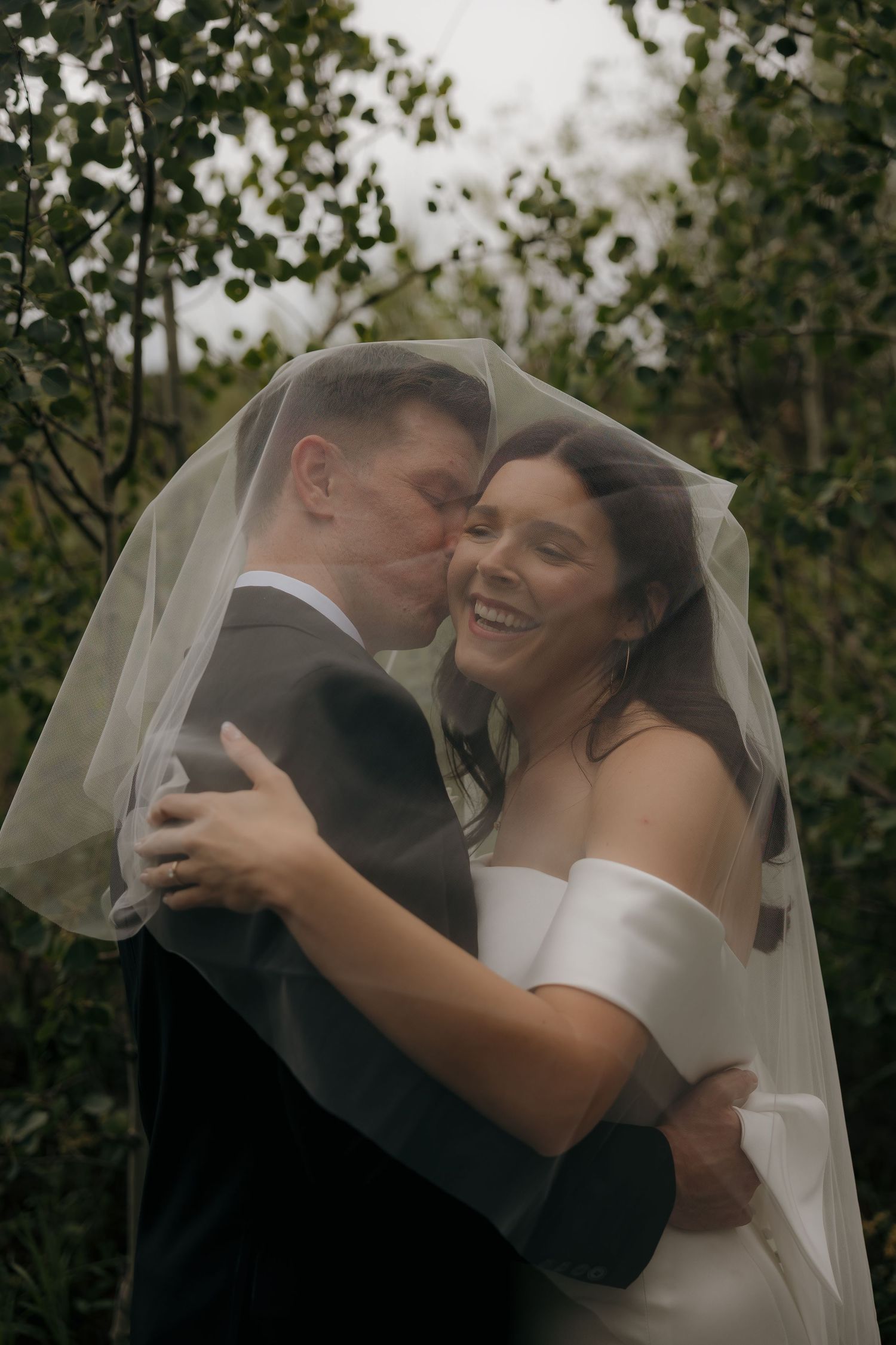 Bride and groom sharing a tender kiss under a sheer veil in lush greenery.