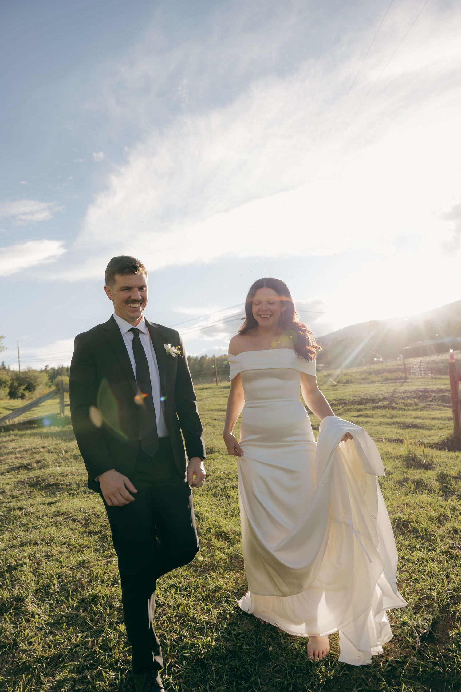 Bride and groom walking hand in hand through green field with golden sunlight glowing behind them.