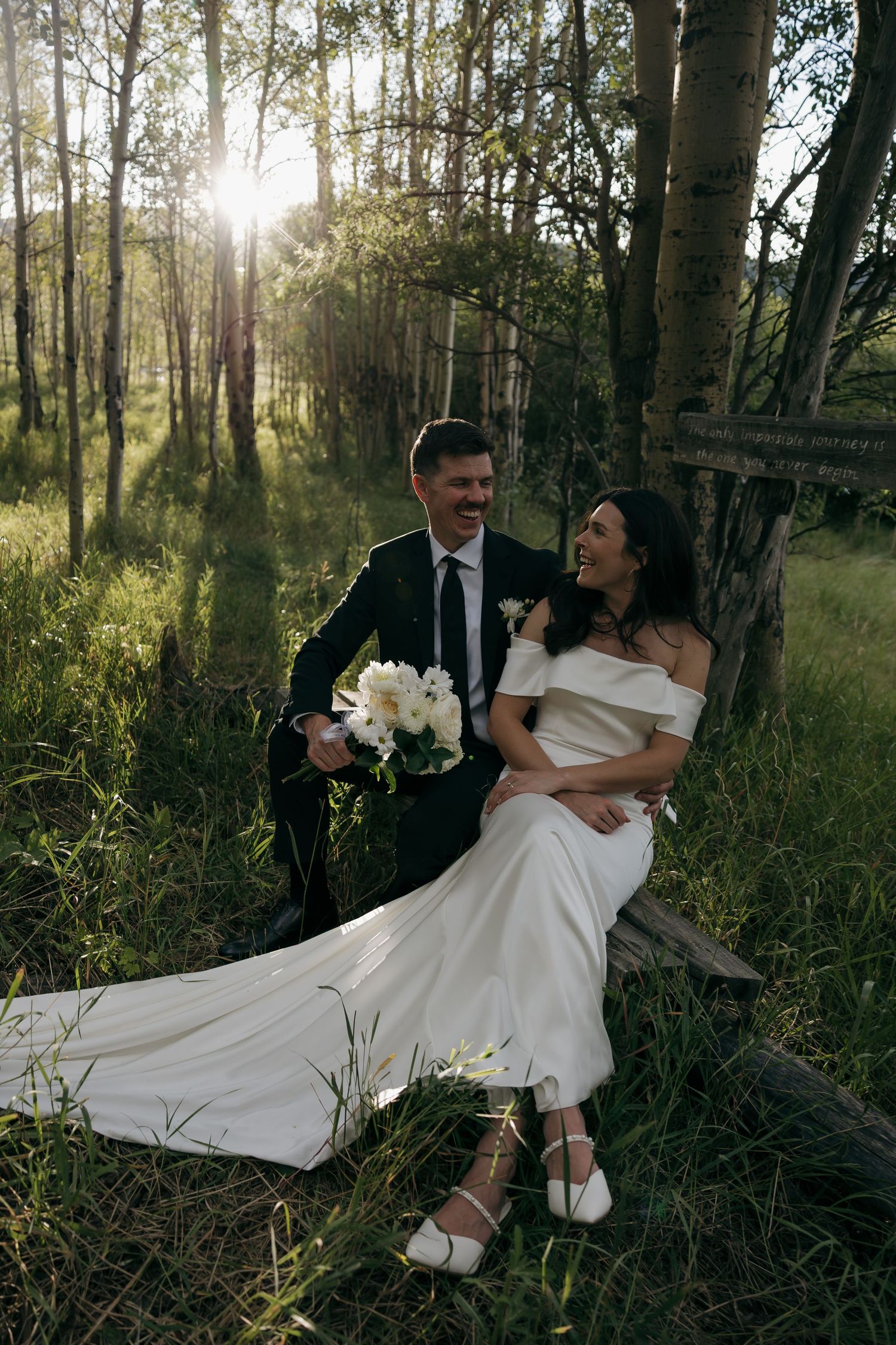 Laughing bride and groom sitting together on a log in a sunlit birch forest during golden hour portraits.