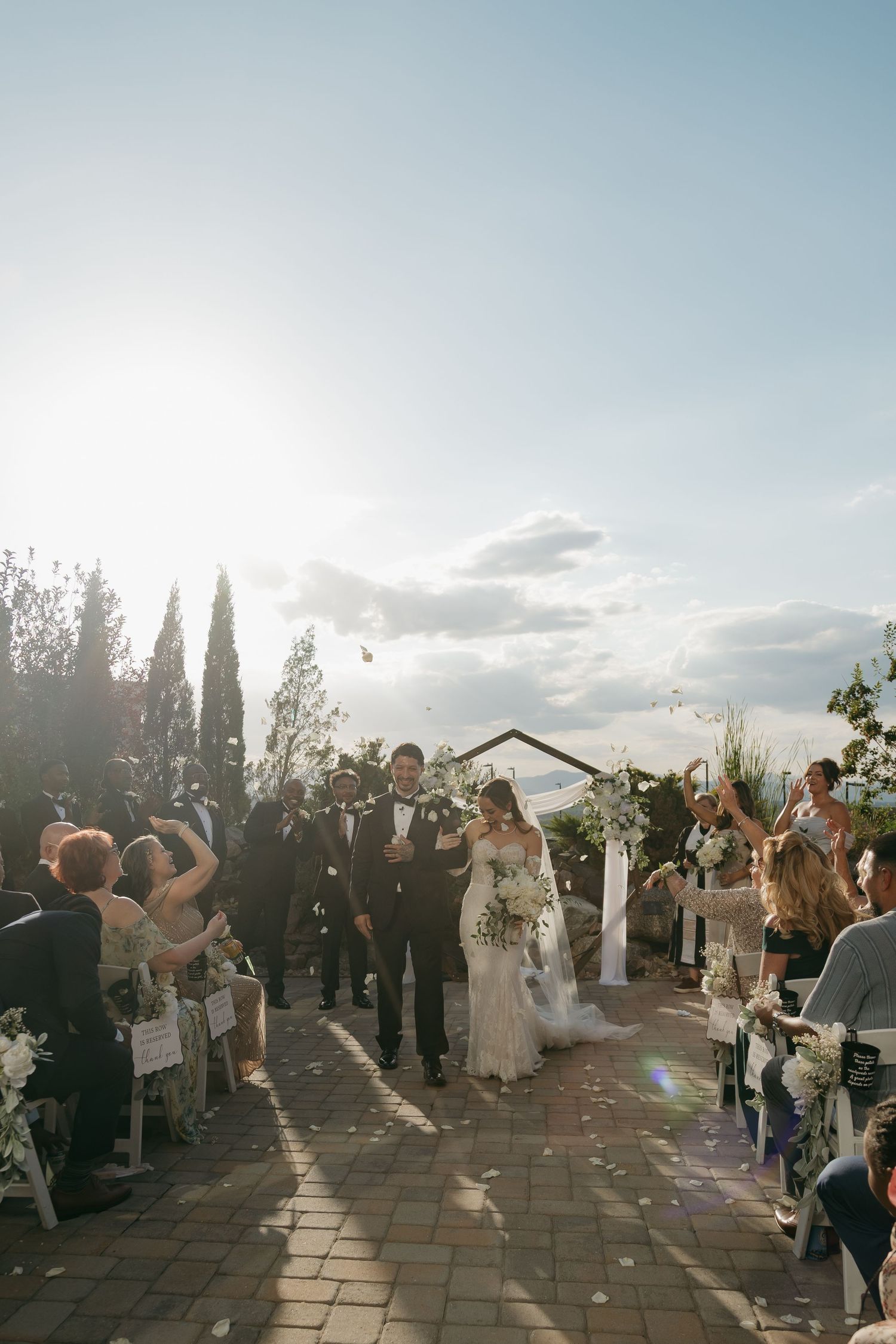 Newlyweds walk down the aisle as guests toss flower petals in a sunlit outdoor ceremony.