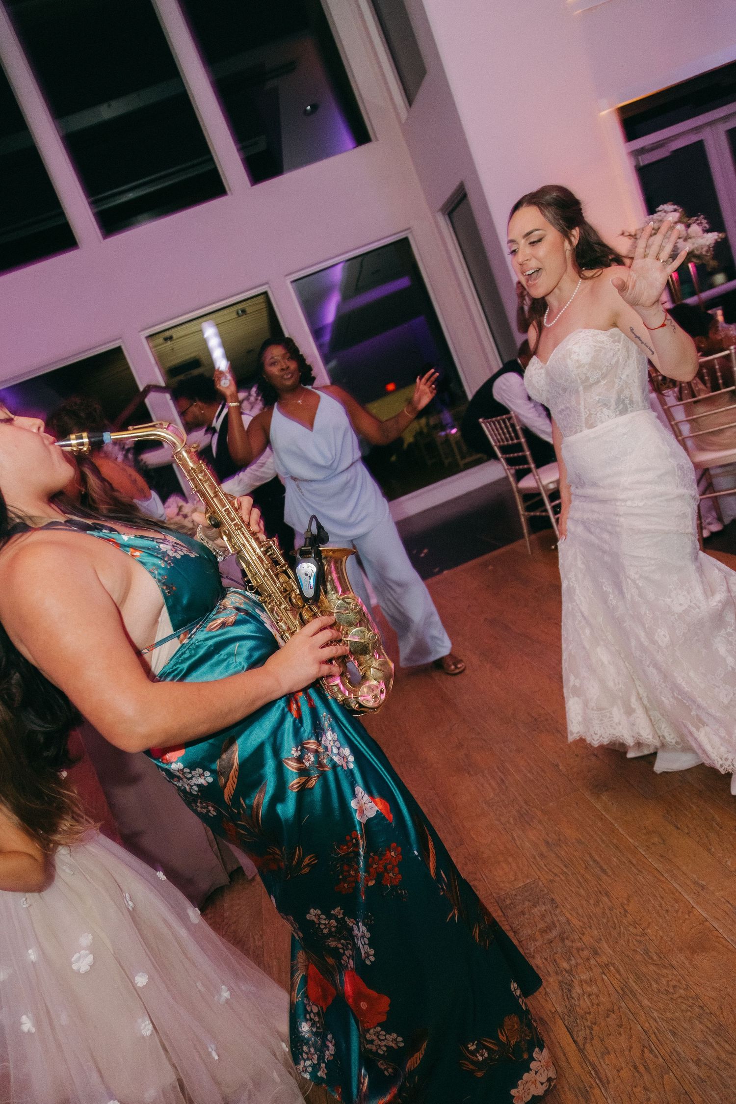 Bride dancing joyfully on wedding reception dance floor while guest plays saxophone in teal gown.