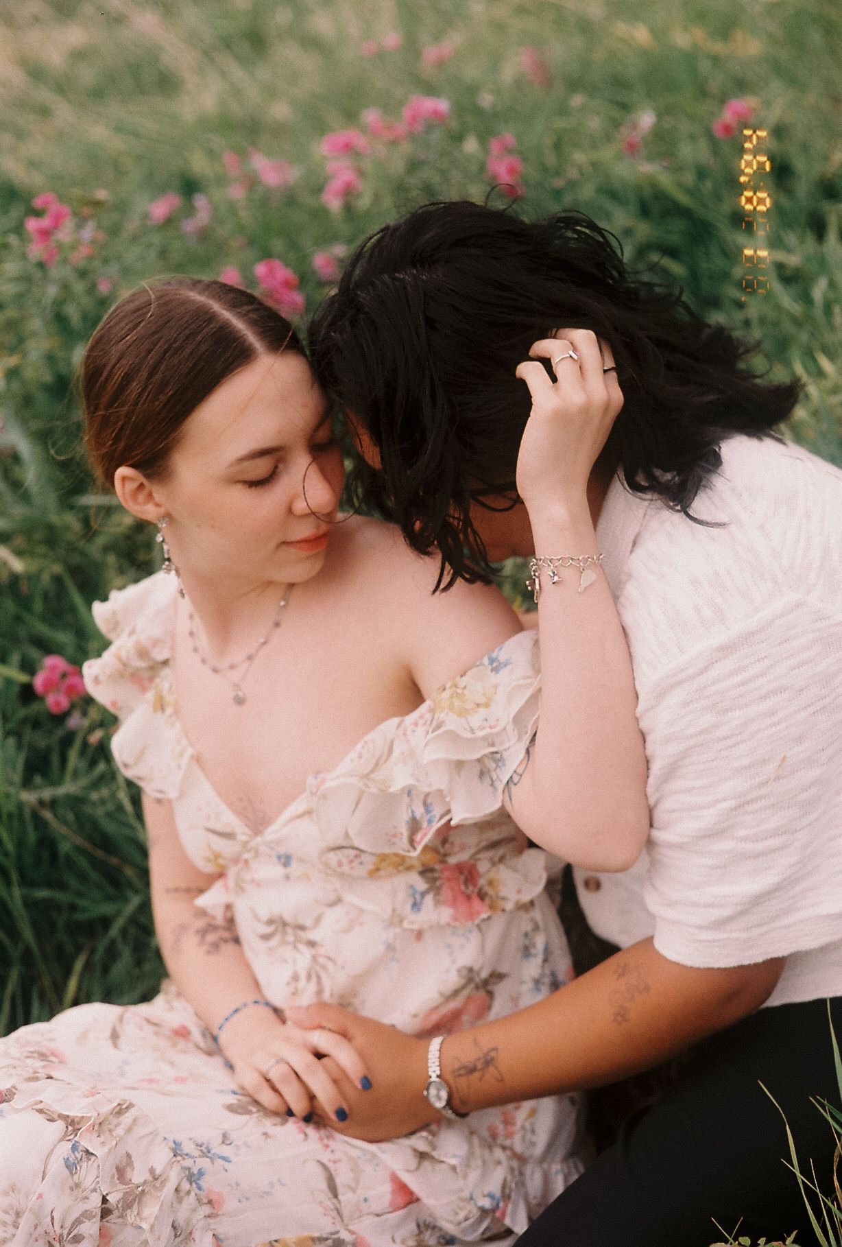 Intimate couple sitting in a flower field, woman in floral dress with partner leaning close.