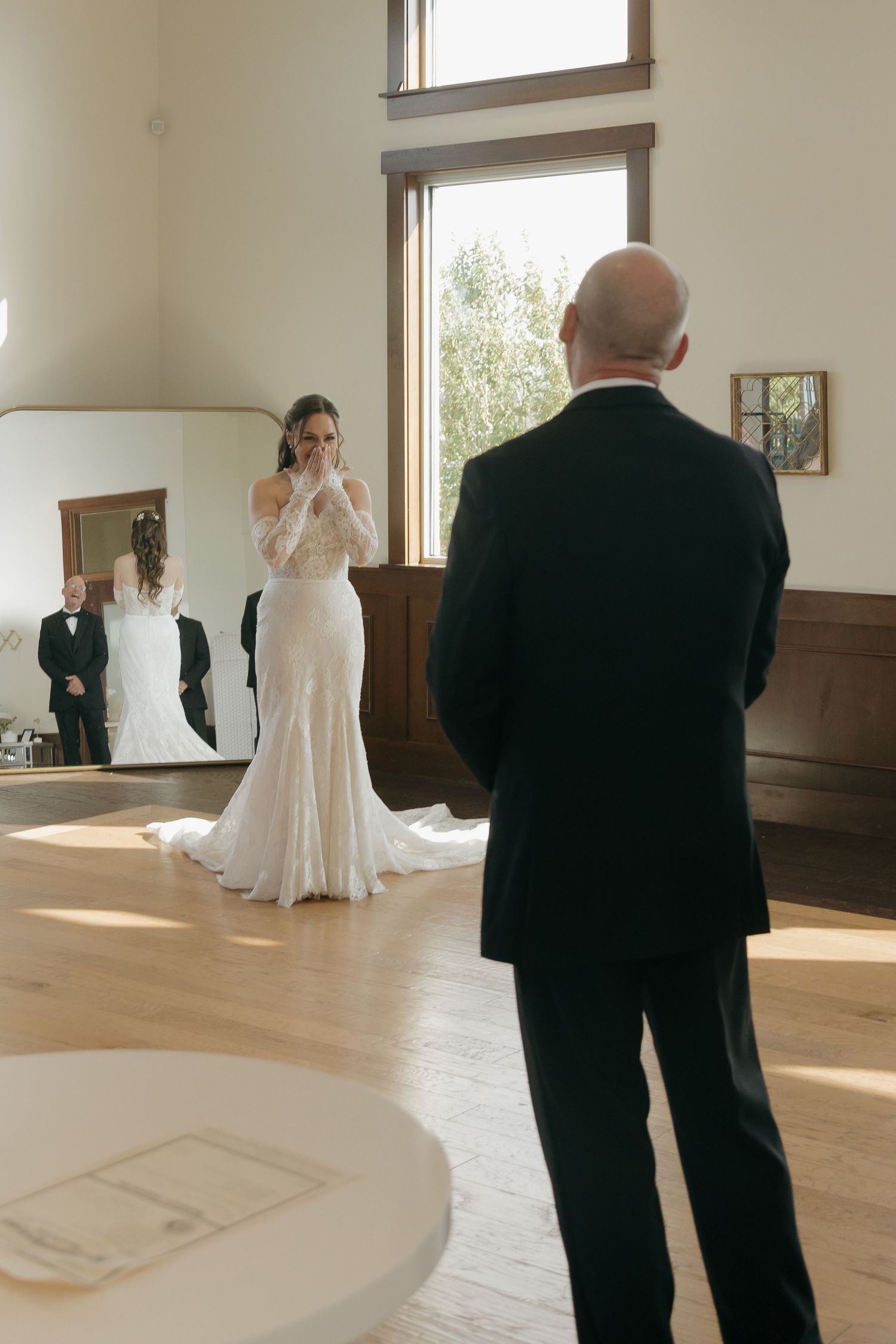 Emotional bride in lace gown covering her face during first look with bald groom indoors.