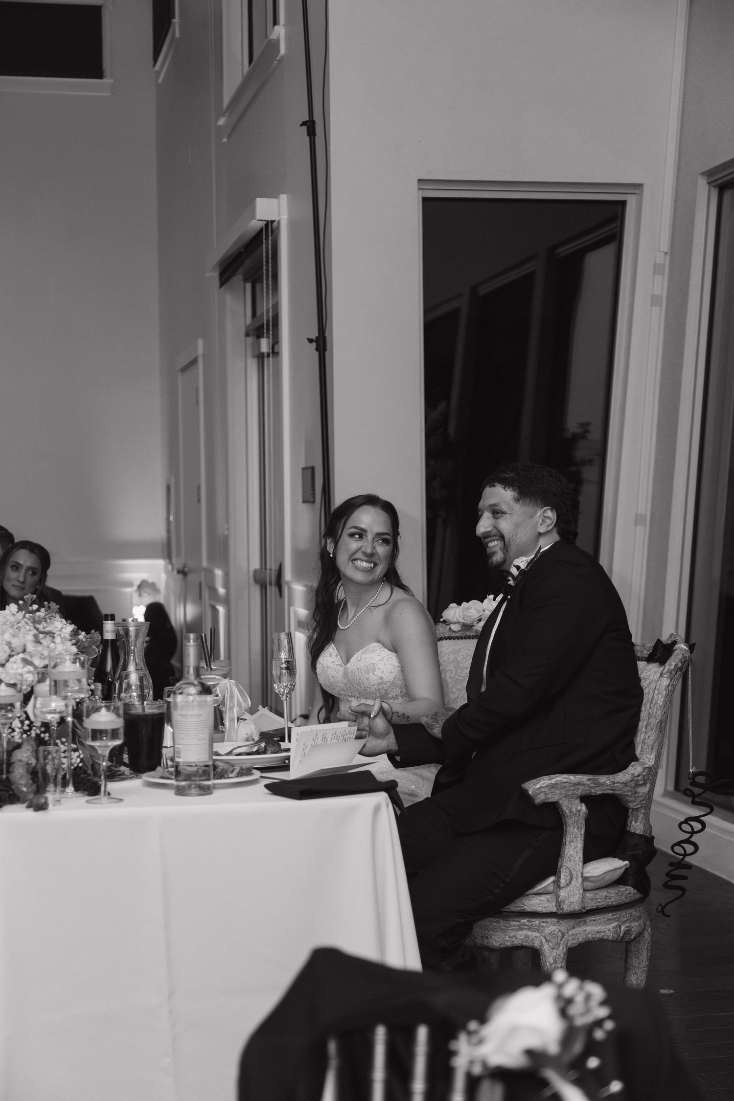 Black and white photo of bride and groom laughing joyfully at sweetheart table during reception.