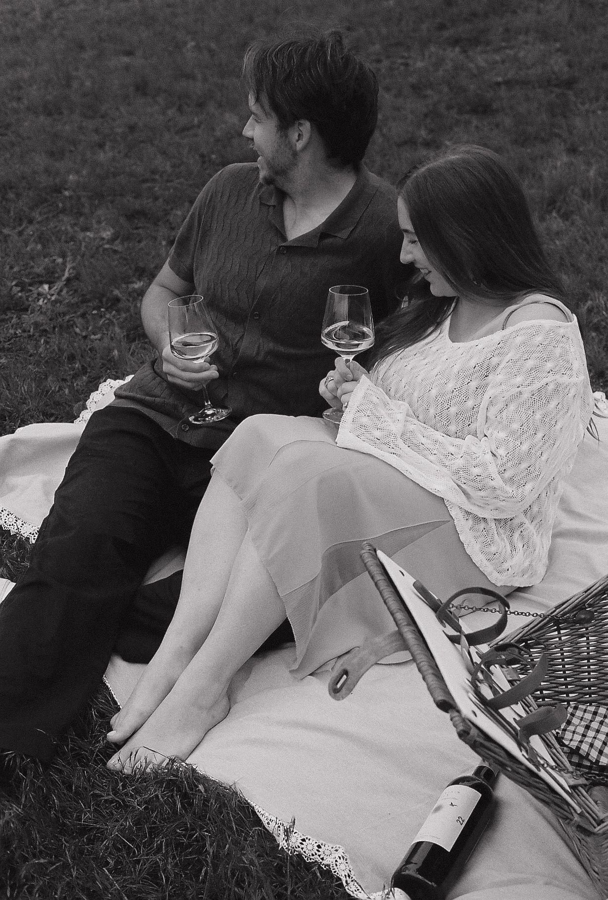 Black and white photo of a couple enjoying wine at a romantic outdoor picnic on a blanket.