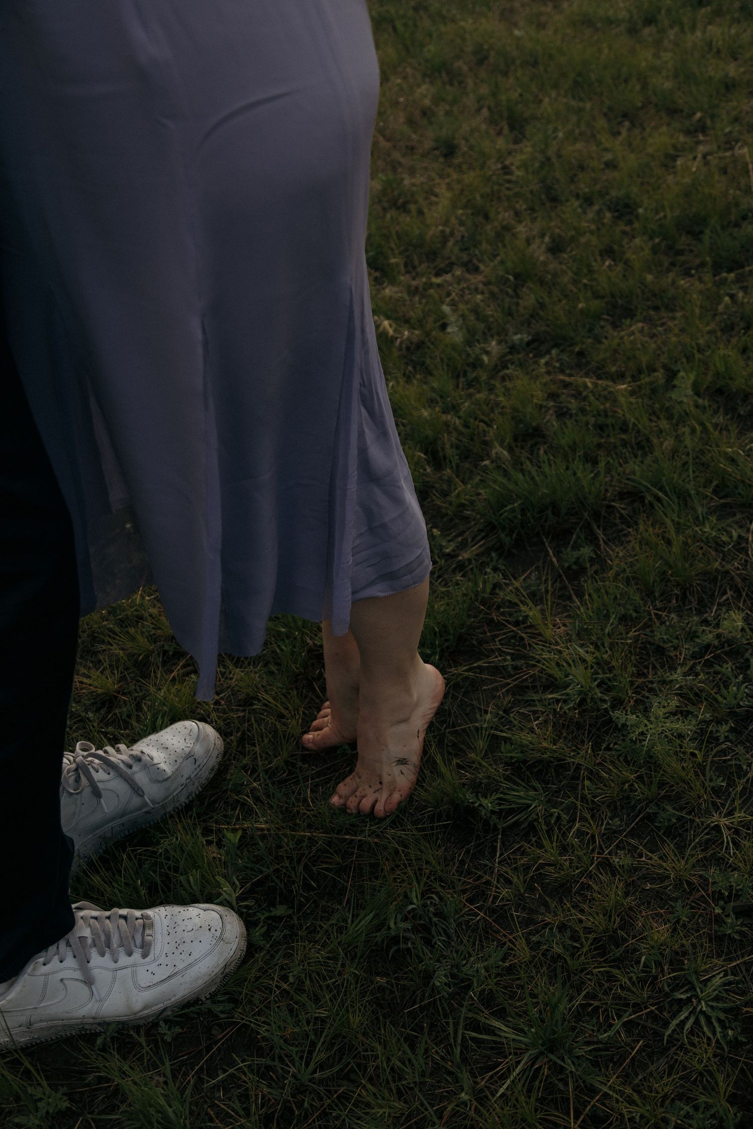Barefoot woman in lavender dress standing on green grass beside man in white sneakers.