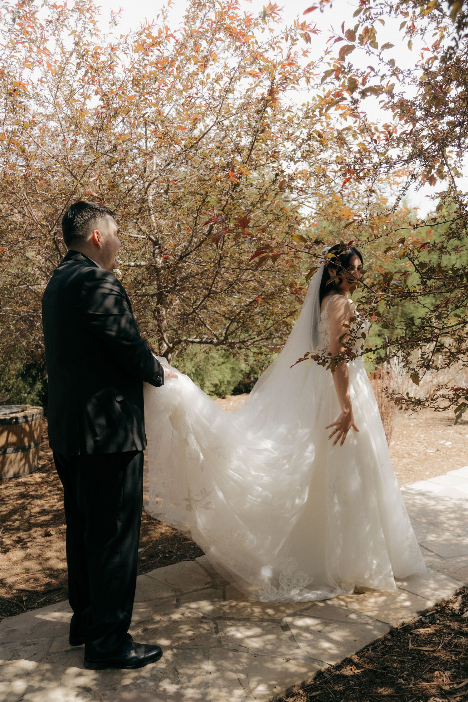 Bride in white gown twirls her flowing dress train outdoors near autumn trees while groom watches.