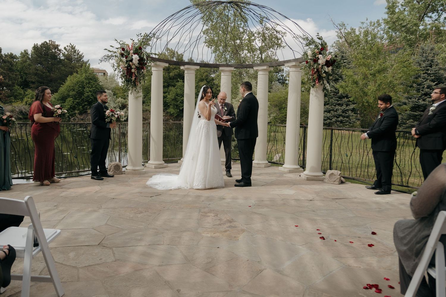 Bride and groom stand at outdoor wedding altar surrounded by white columns and lush greenery.