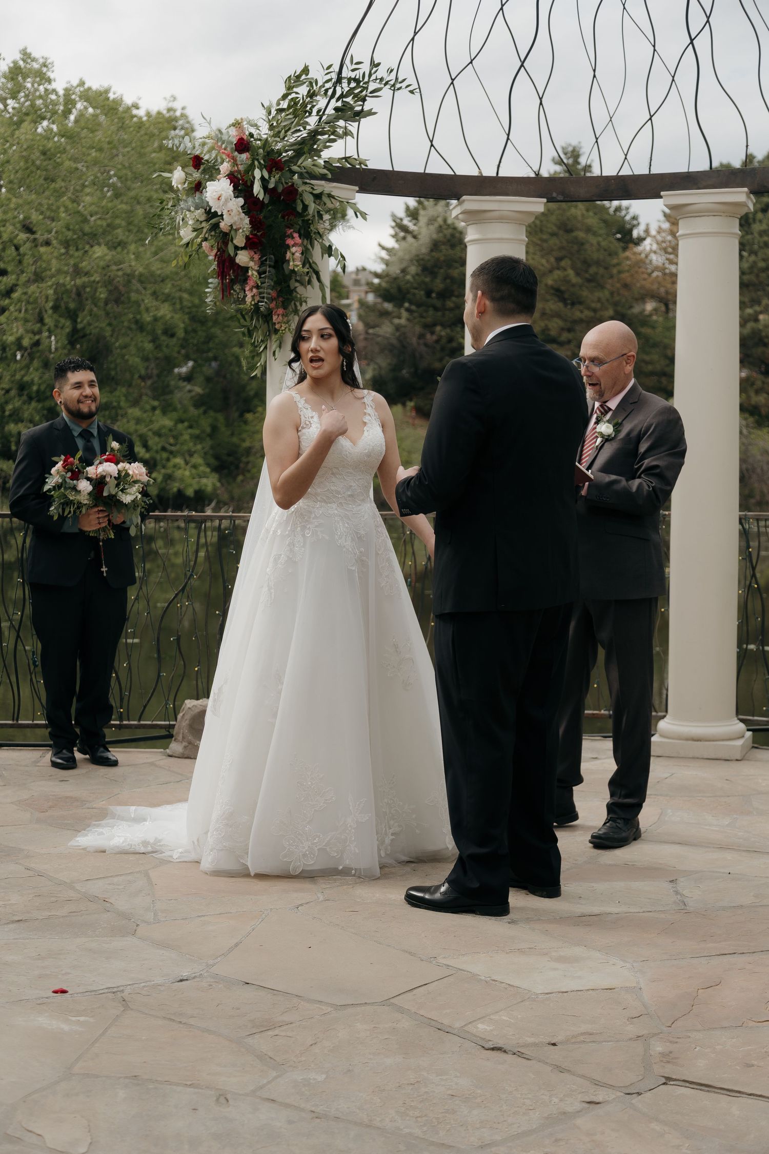 Bride in lace ball gown recites wedding vows to groom at elegant outdoor ceremony beneath floral archway.
