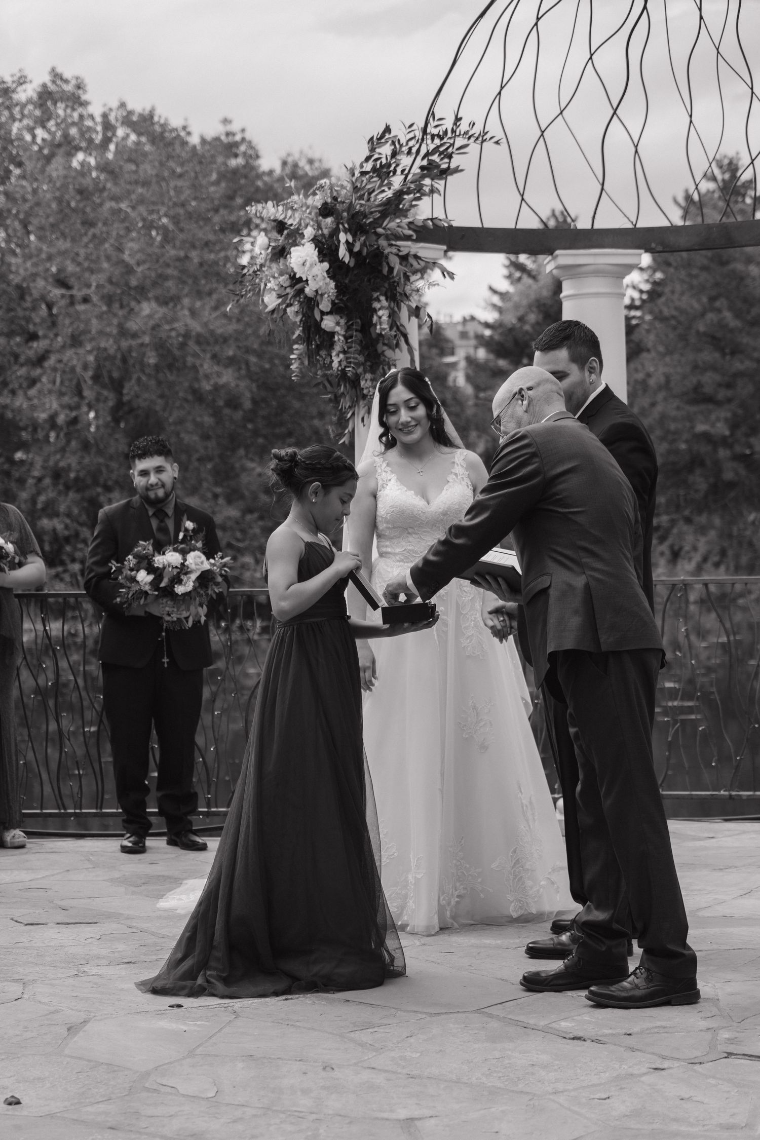 Black and white wedding ceremony photo of bride receiving ring from officiant with bridesmaid and guests watching.
