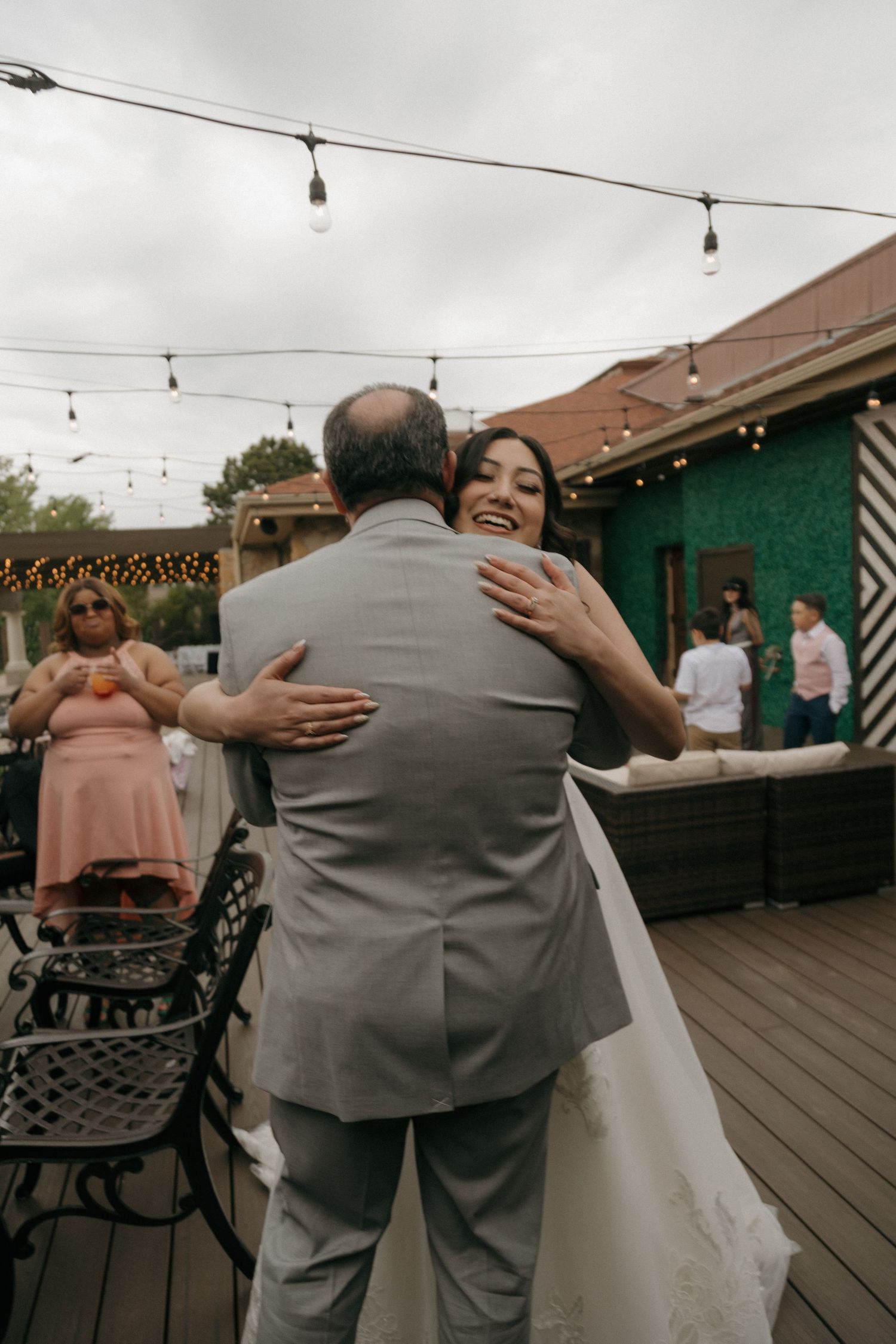 Smiling bride in white gown shares a warm hug on a festive outdoor deck adorned with string lights at her wedding reception.