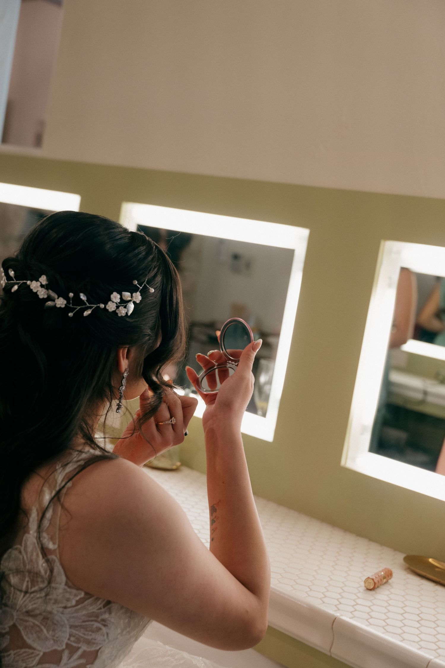 Bride applying makeup at a vanity mirror with Hollywood lights, wearing a floral hair crown on her wedding day.