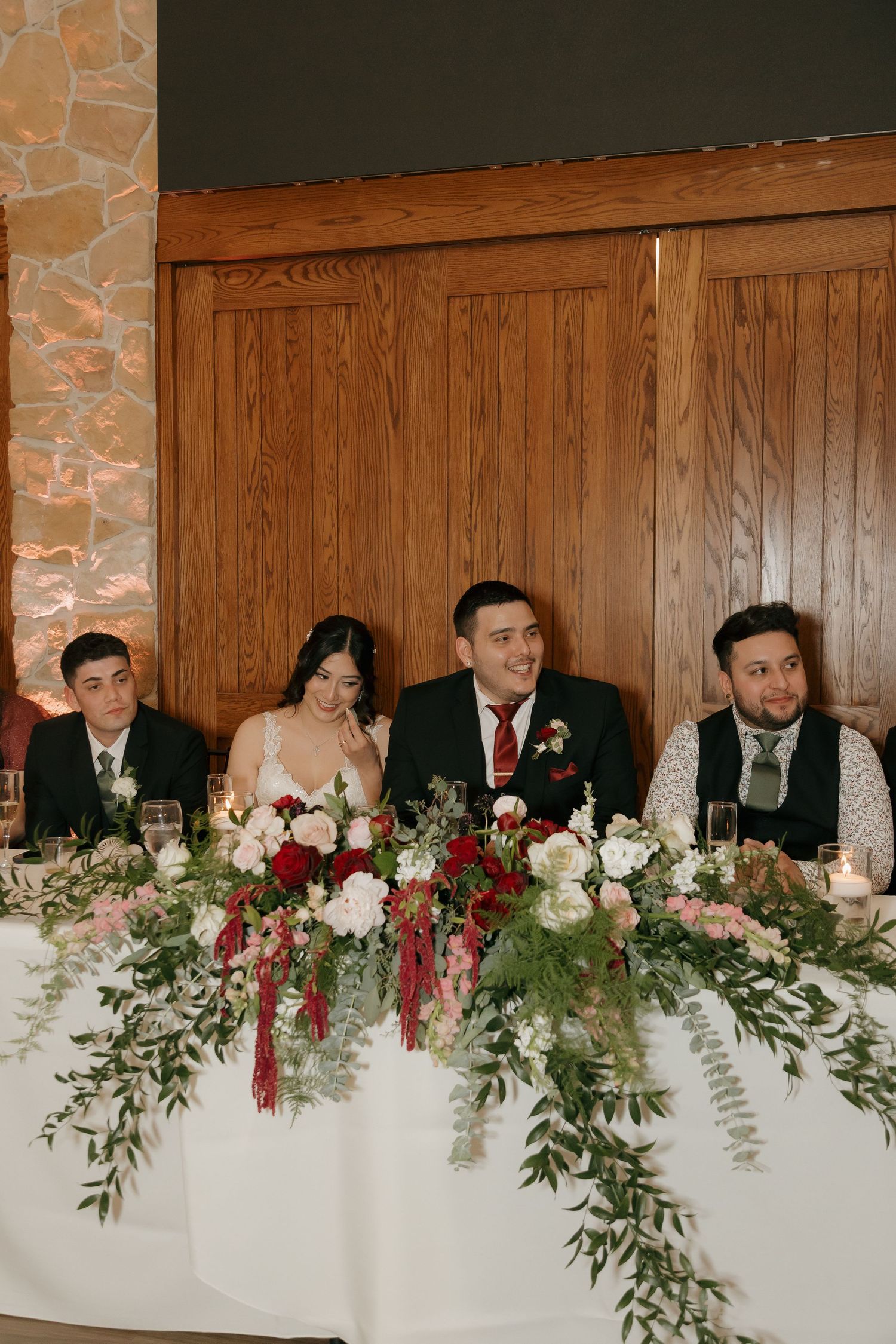 Bride and groom smile at floral-adorned head table during wedding reception with wedding party.
