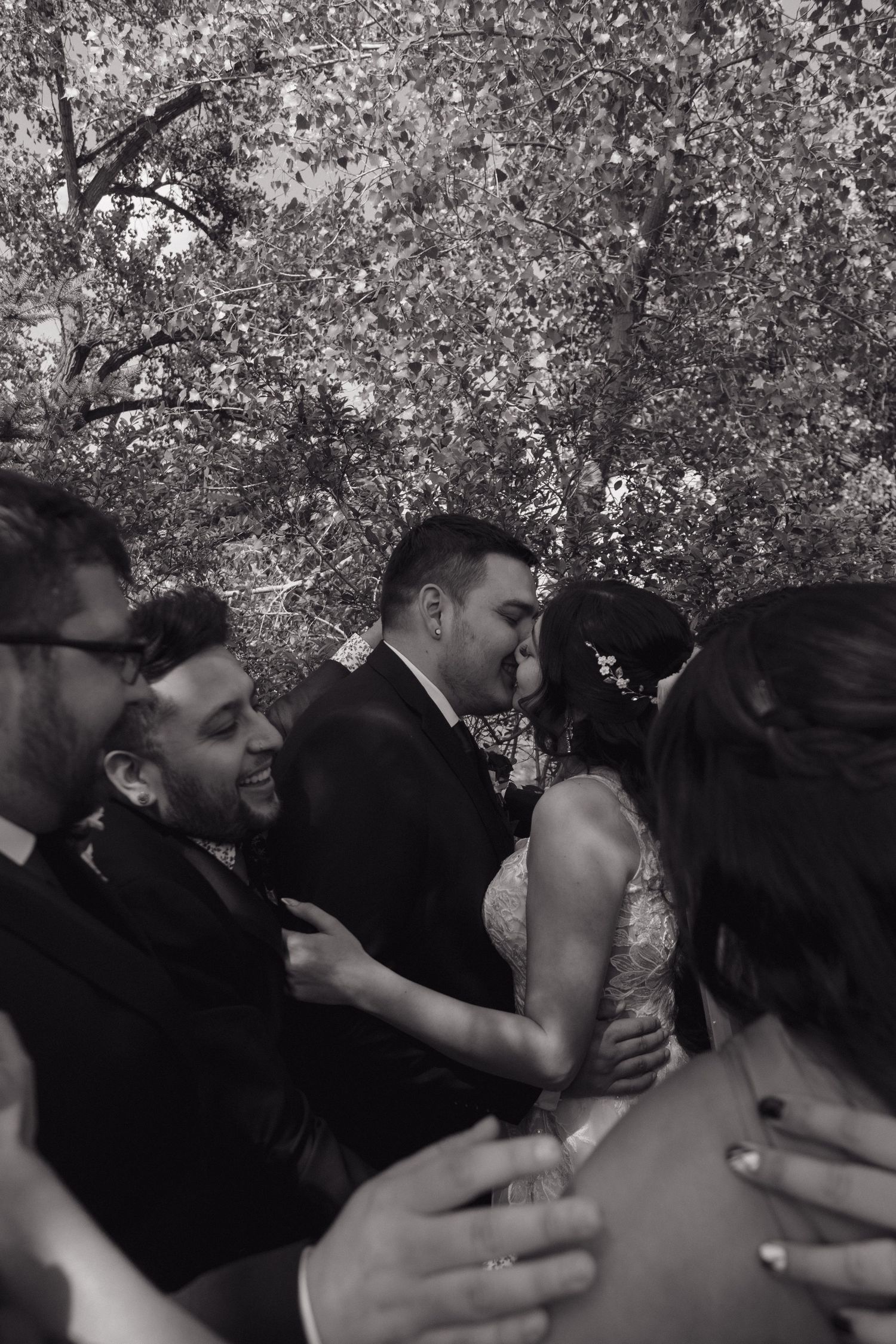 Black and white photo of bride and groom sharing a kiss surrounded by smiling wedding party members.