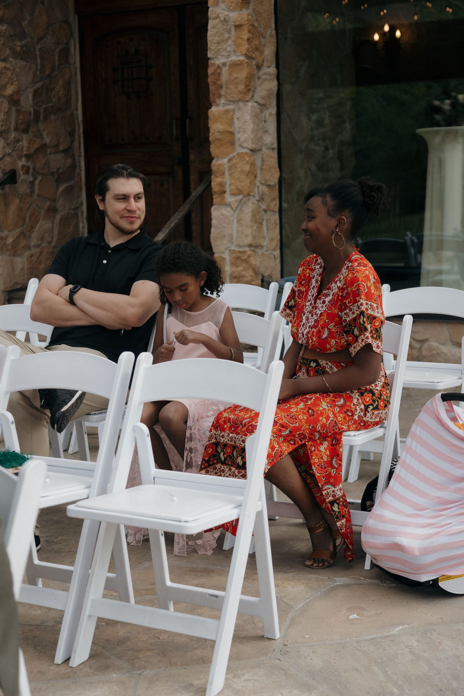 Wedding guests including a man, young girl, and woman in floral dress seated in white chairs at ceremony.
