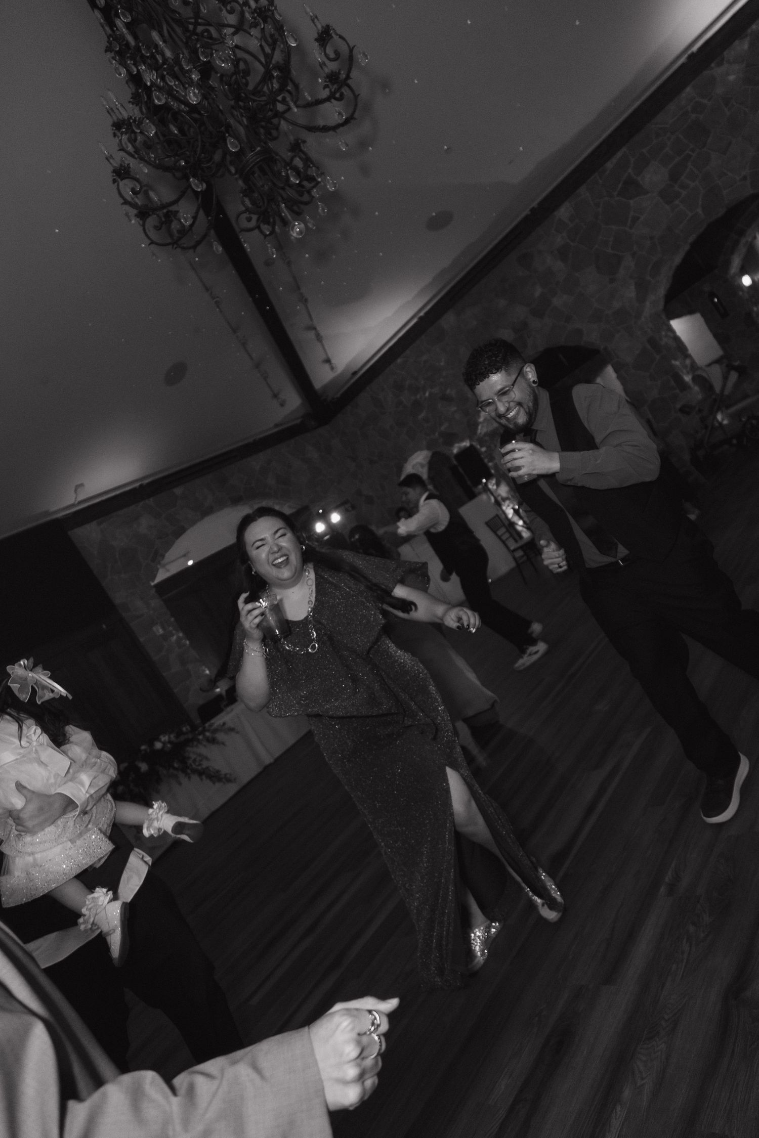 Guests laugh and dance at wedding reception in dramatic tilted black and white photograph.