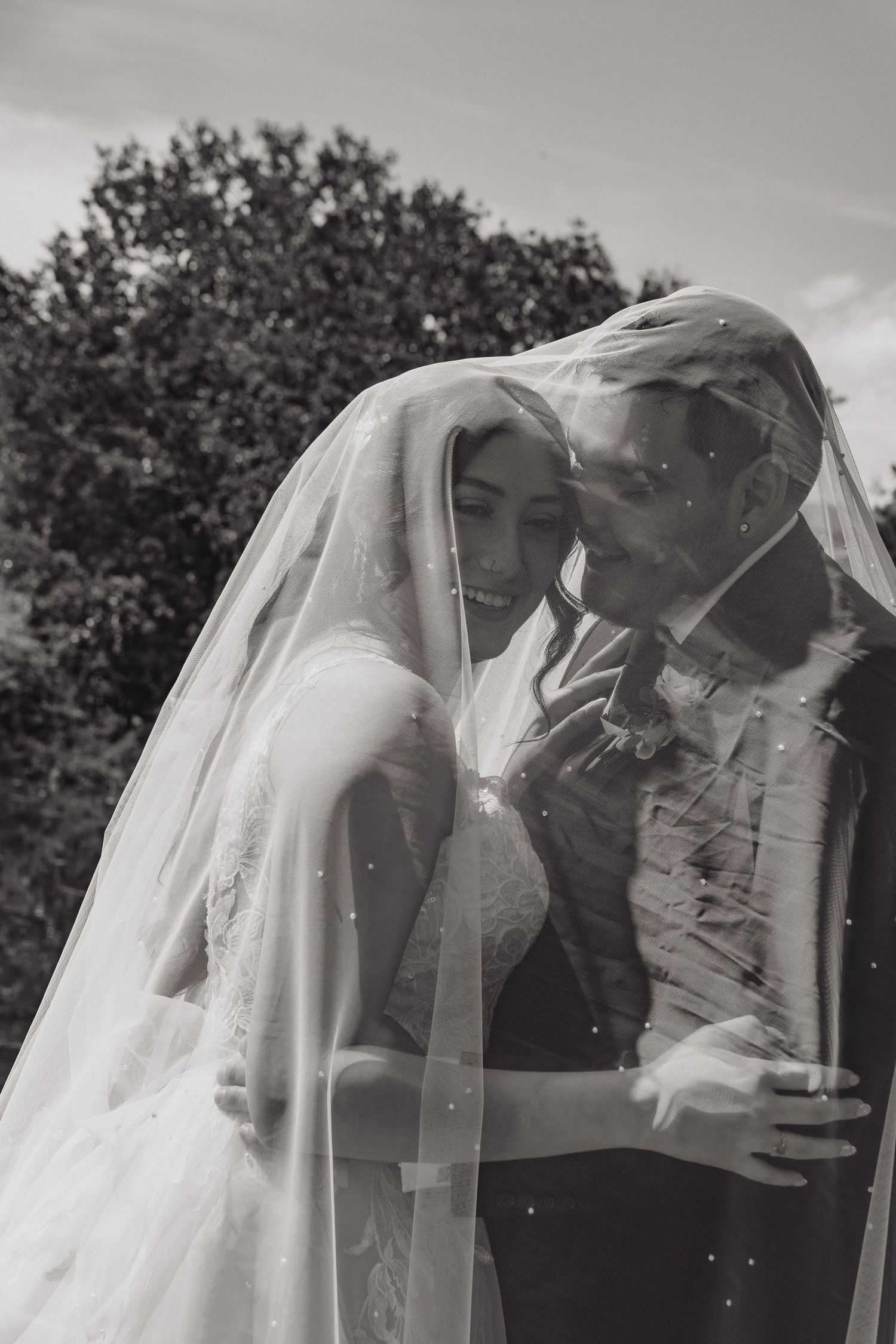 Black and white photo of bride and groom embracing tenderly under a flowing veil outdoors.