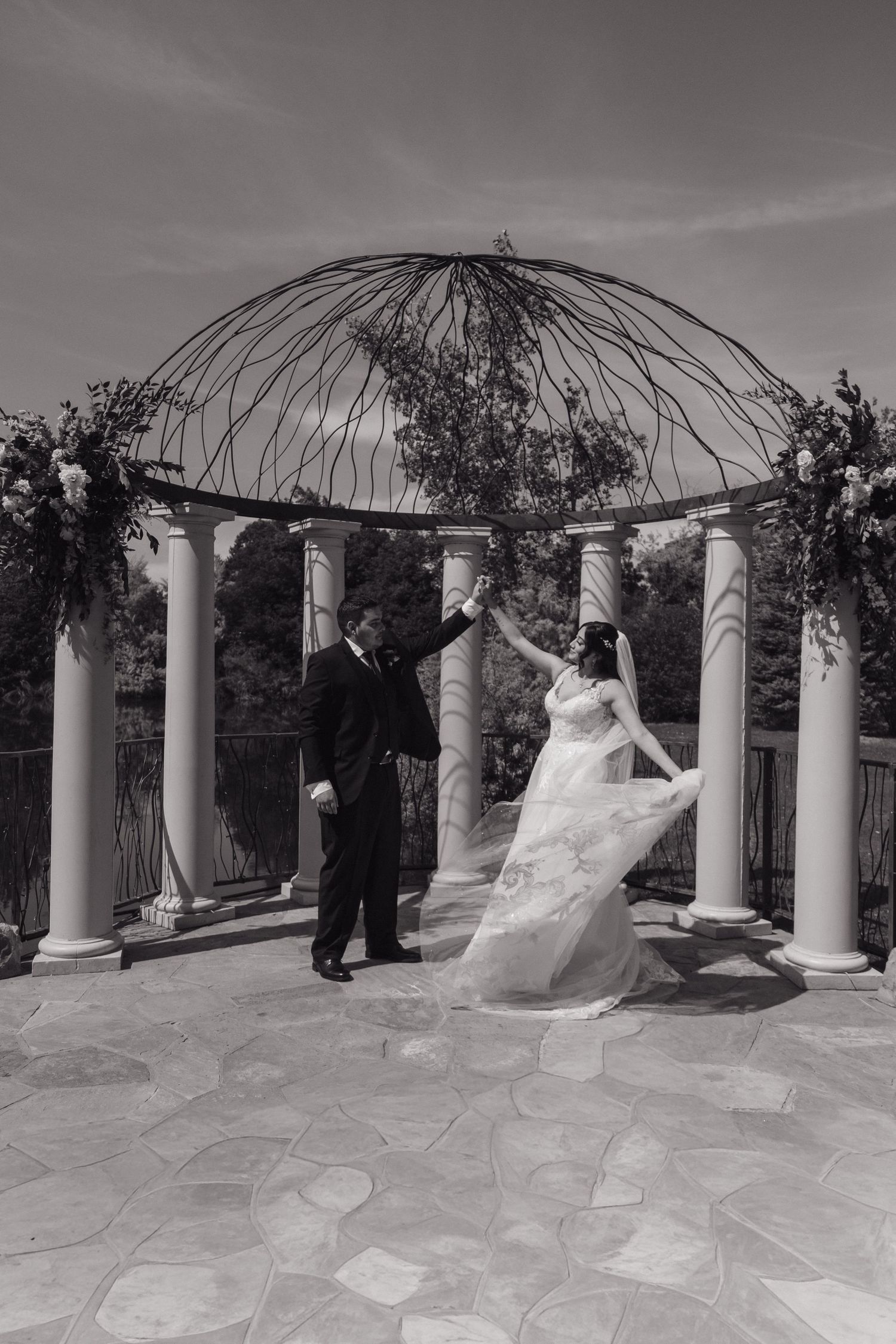 Black and white photo of bride and groom dancing under ornate gazebo with floral arrangements and columns.