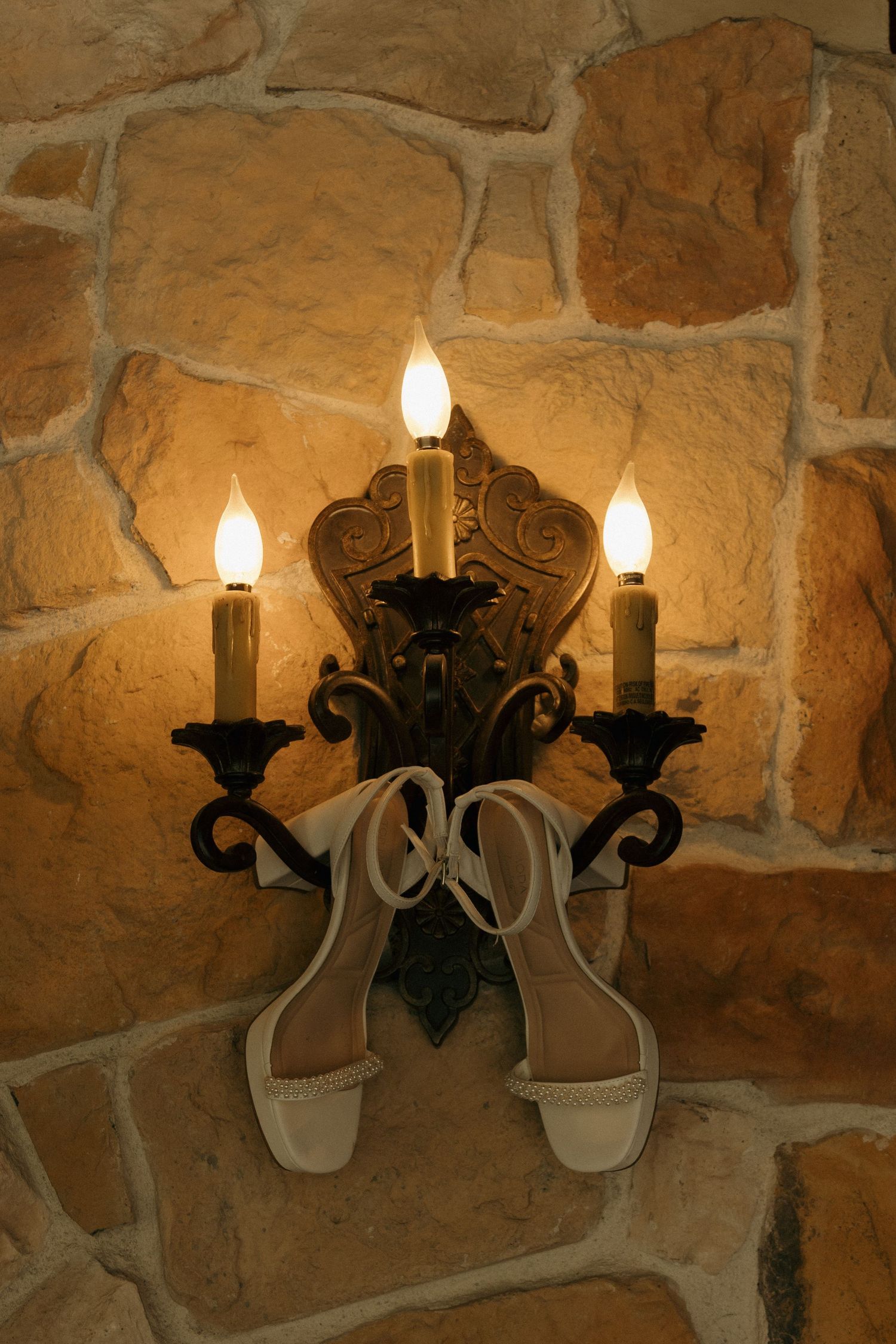 White bridal heels hanging on an ornate gold candelabra wall sconce against a rustic stone wall.