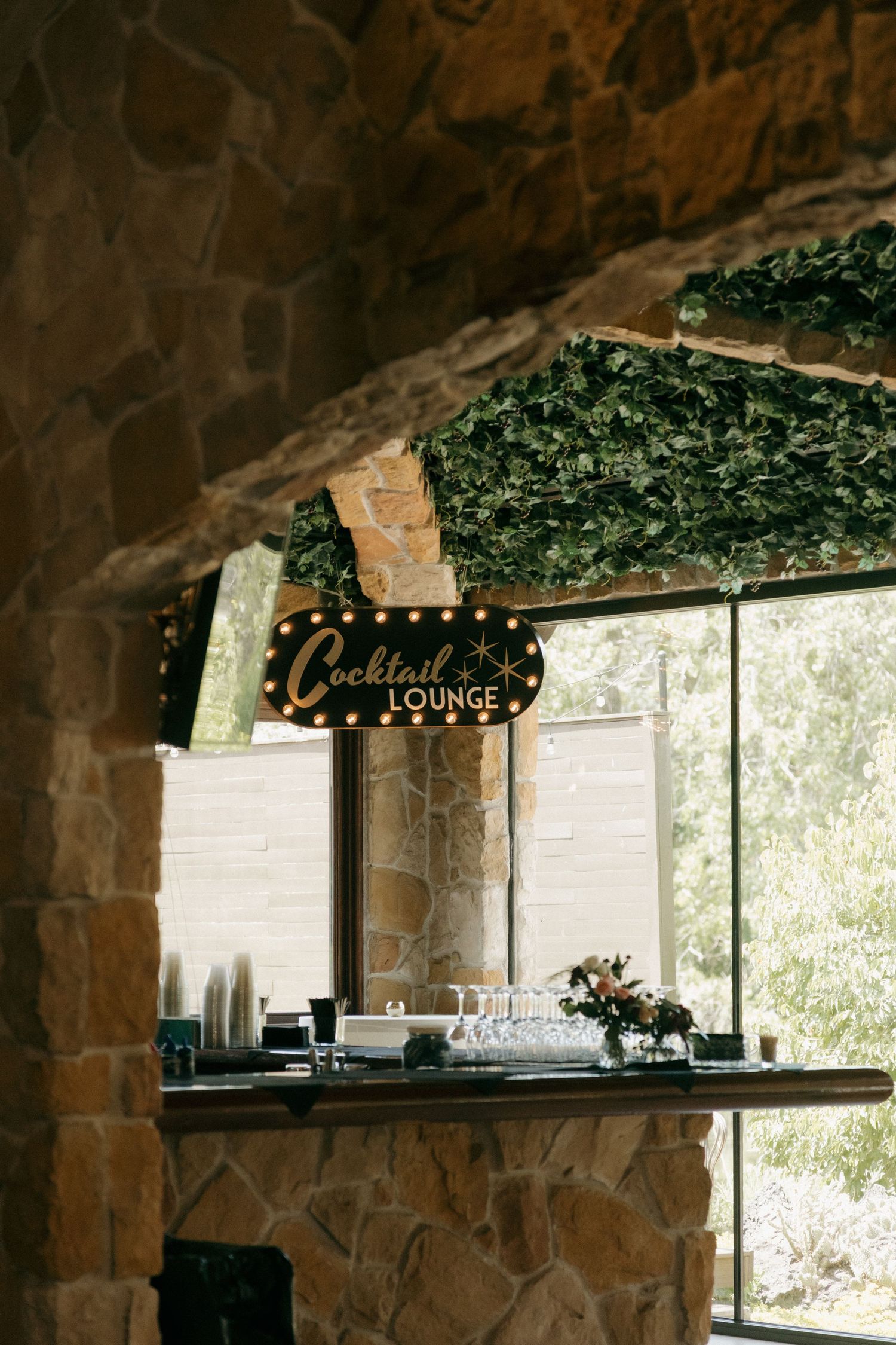 A vintage marquee Cocktail Lounge sign displayed above a stone bar decorated with greenery at a wedding venue.