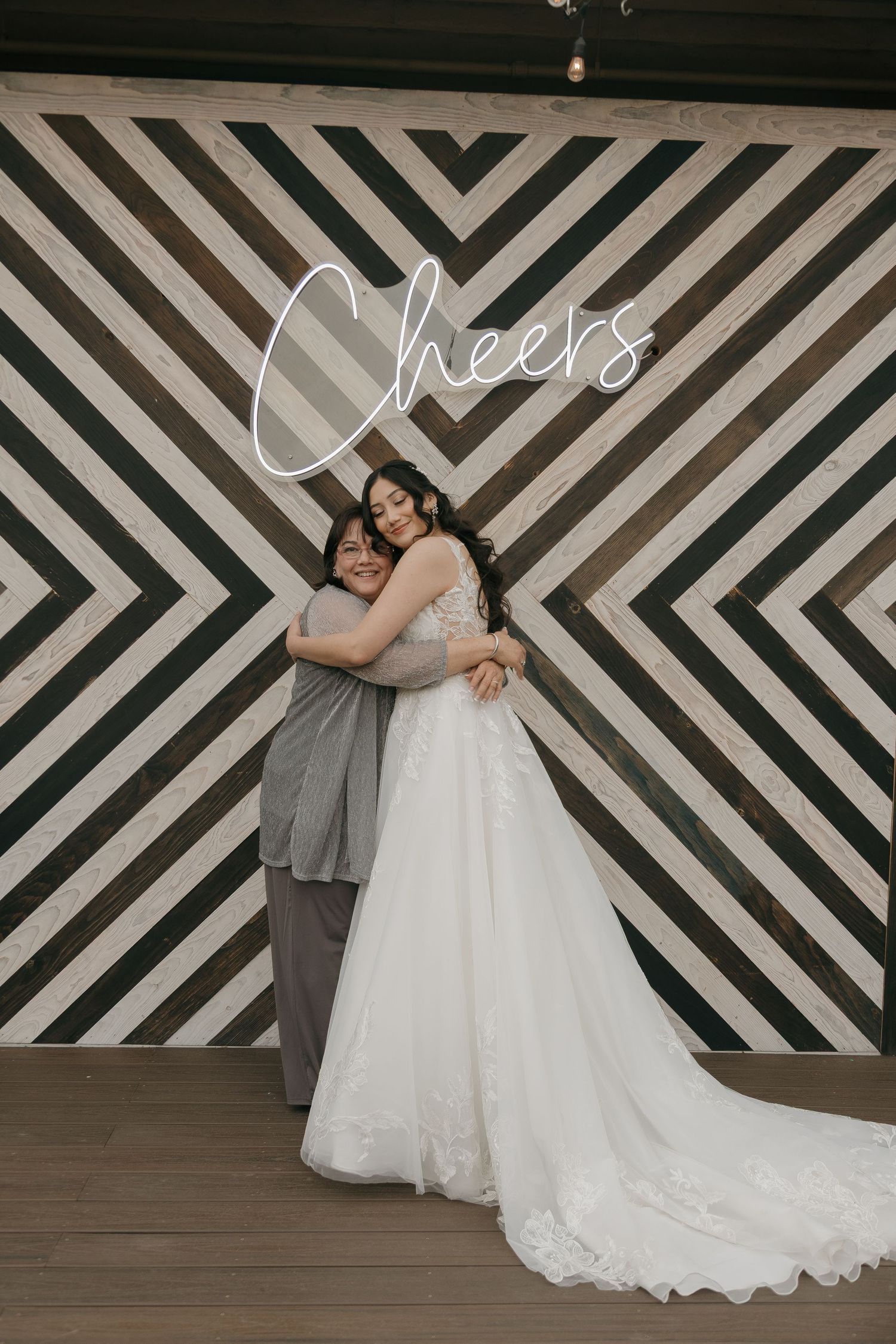 Bride in lace wedding gown embracing a guest in front of a geometric striped Cheers neon sign backdrop.