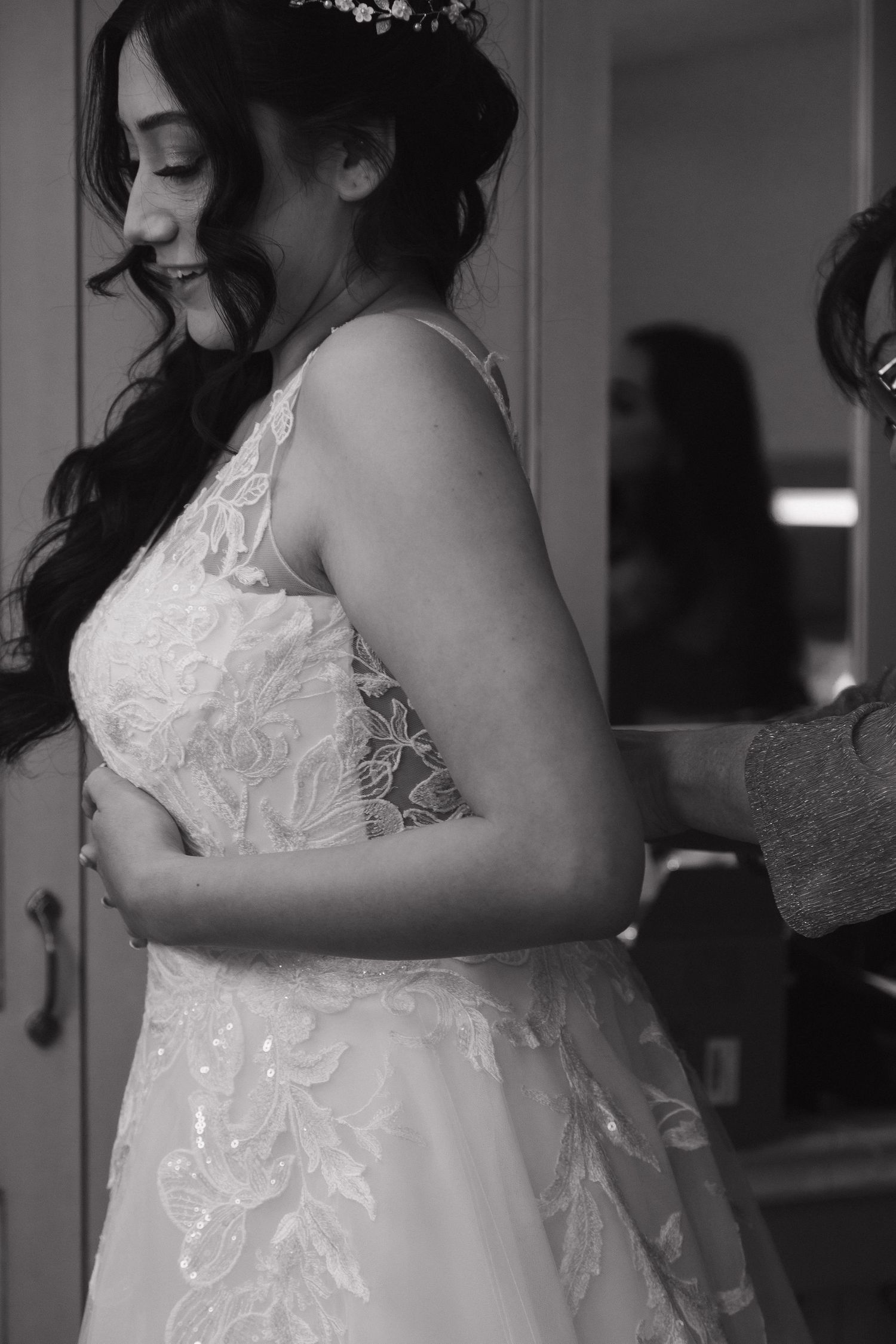 Black and white photo of a bride in lace wedding gown smiling while being helped into her dress.