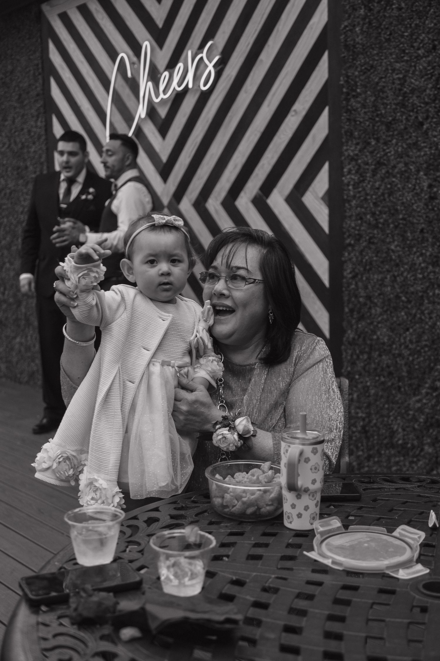 Grandmother joyfully holds a baby girl in white dress at a wedding reception in black and white photo.