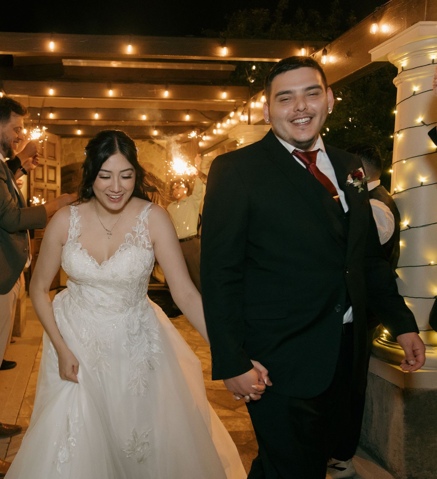 Newlyweds holding hands and smiling during sparkler exit at night wedding celebration outdoors.