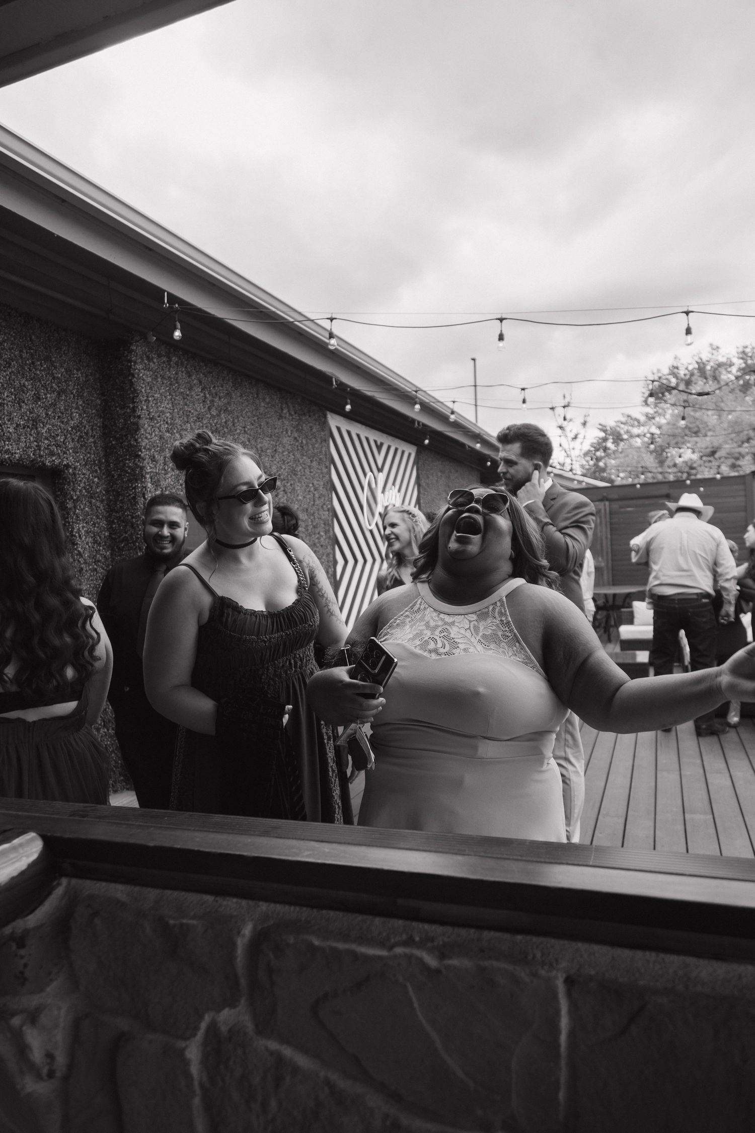 Wedding guests laugh and celebrate outdoors on a patio decorated with string lights in black and white photo.