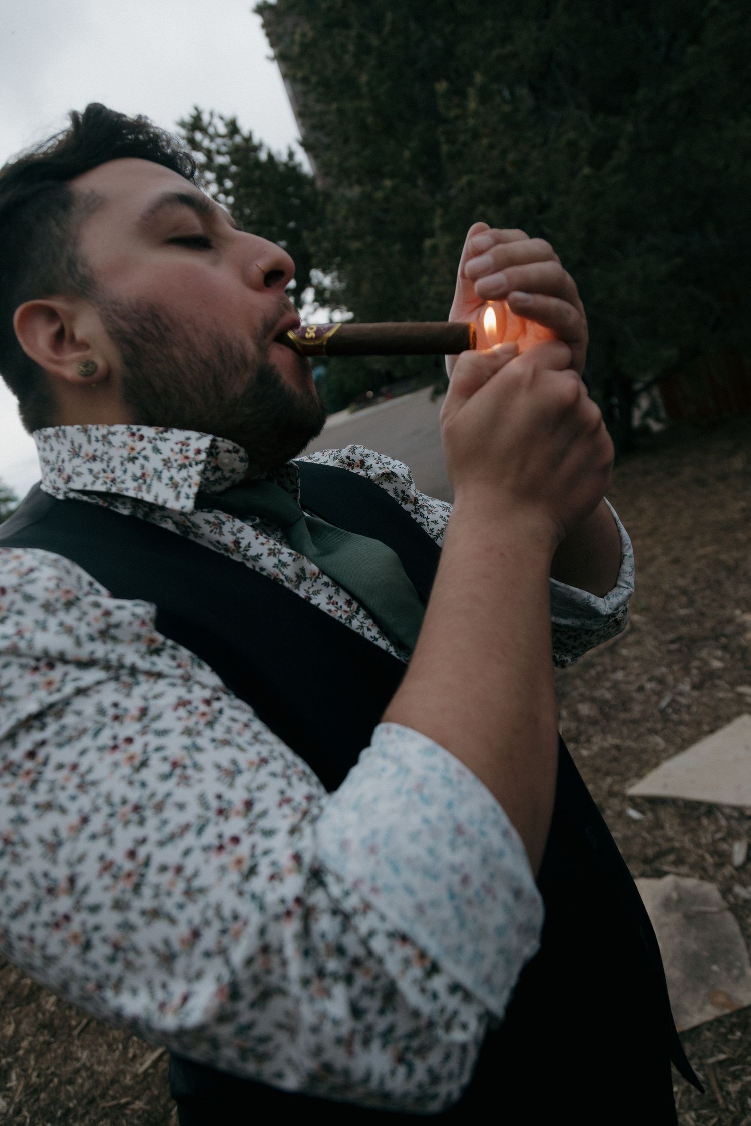 Groomsman in floral shirt and vest lighting a cigar outdoors at wedding reception.