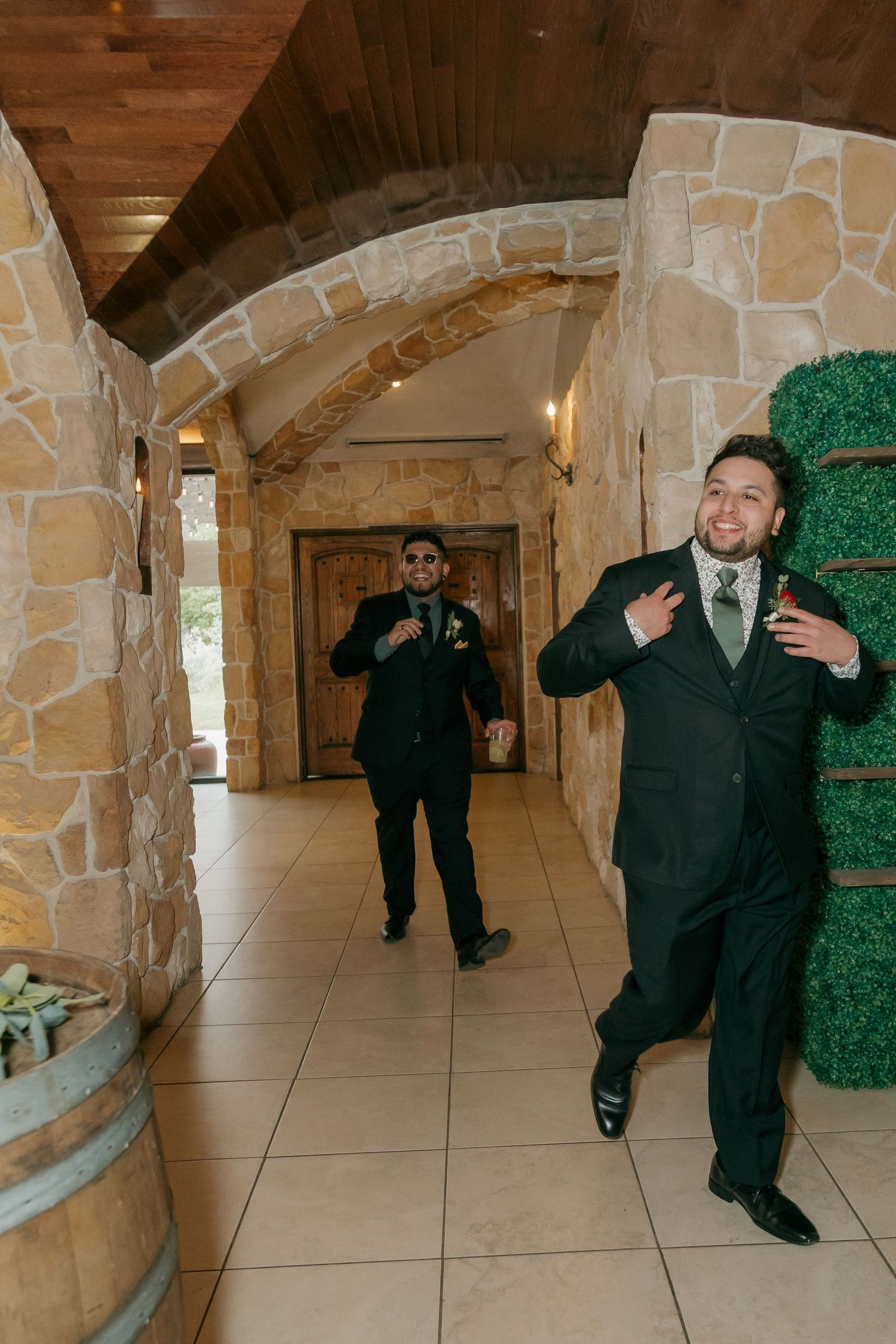 Two groomsmen in dark suits walk energetically through a stone archway corridor at a wedding venue.