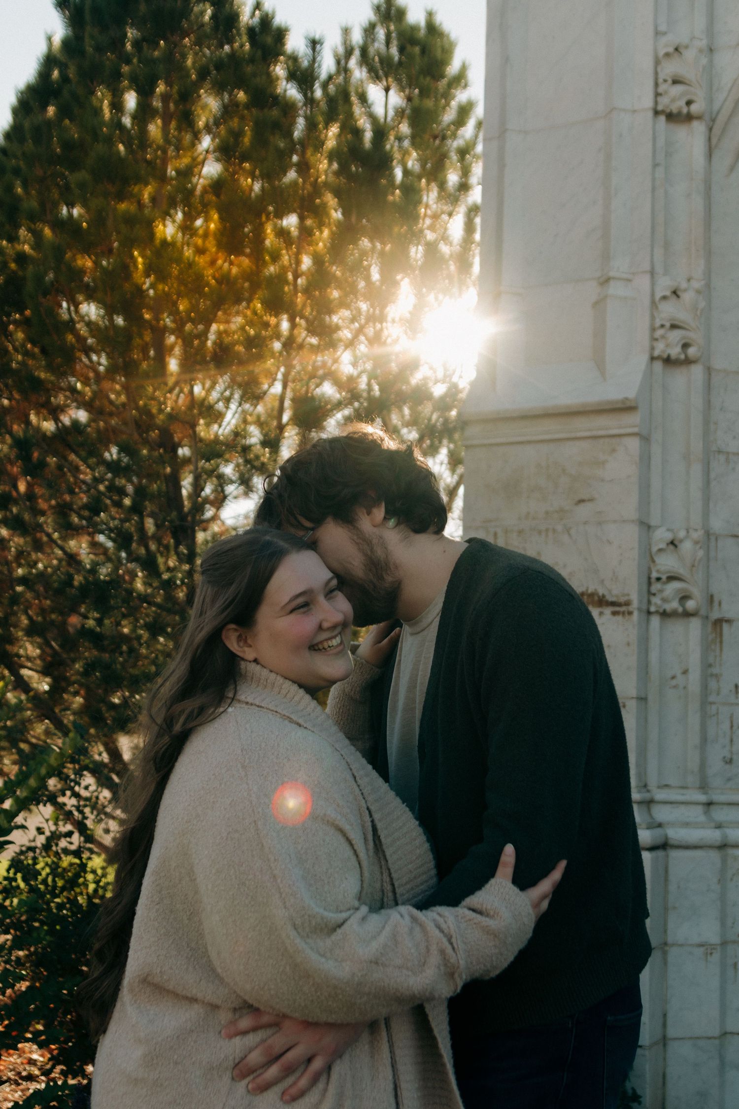 A couple shares a tender embrace at golden hour, with sunlight streaming through autumn trees beside a white stone column.