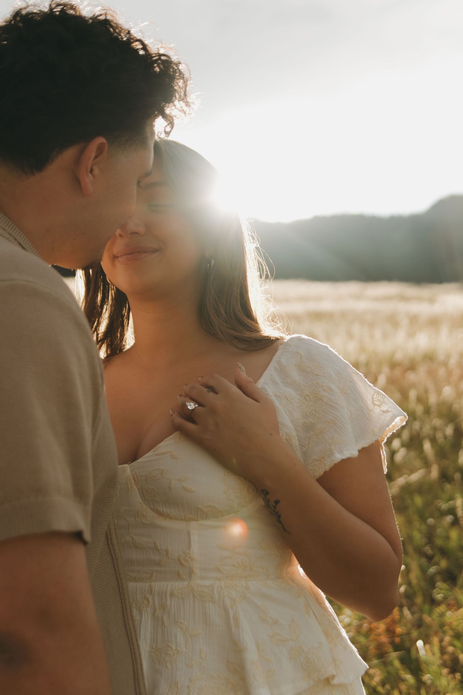 Couple embracing in golden field at sunset, woman in white dress smiling as man kisses her temple.