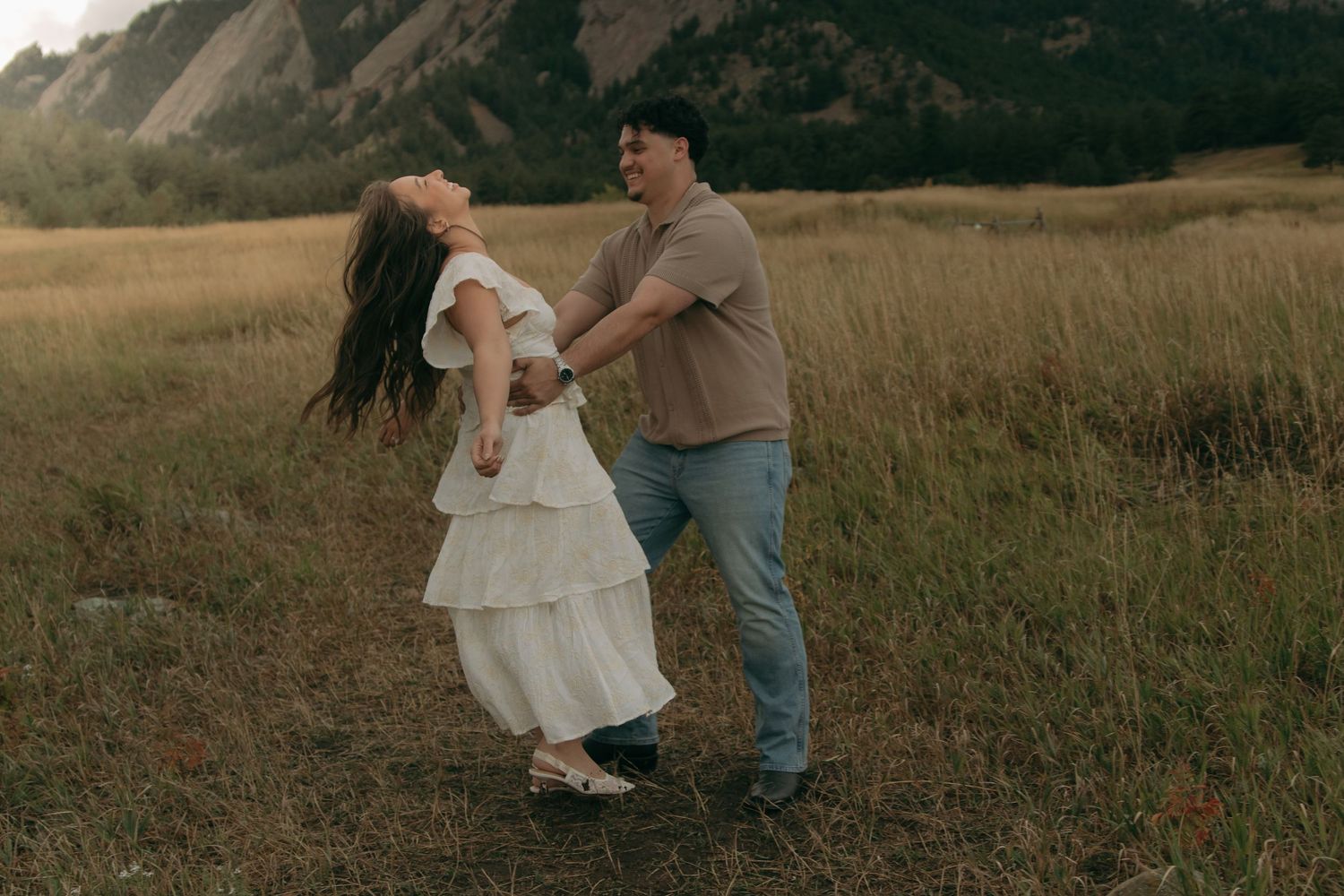A joyful couple dances and spins together in a golden meadow with rocky mountains behind them.